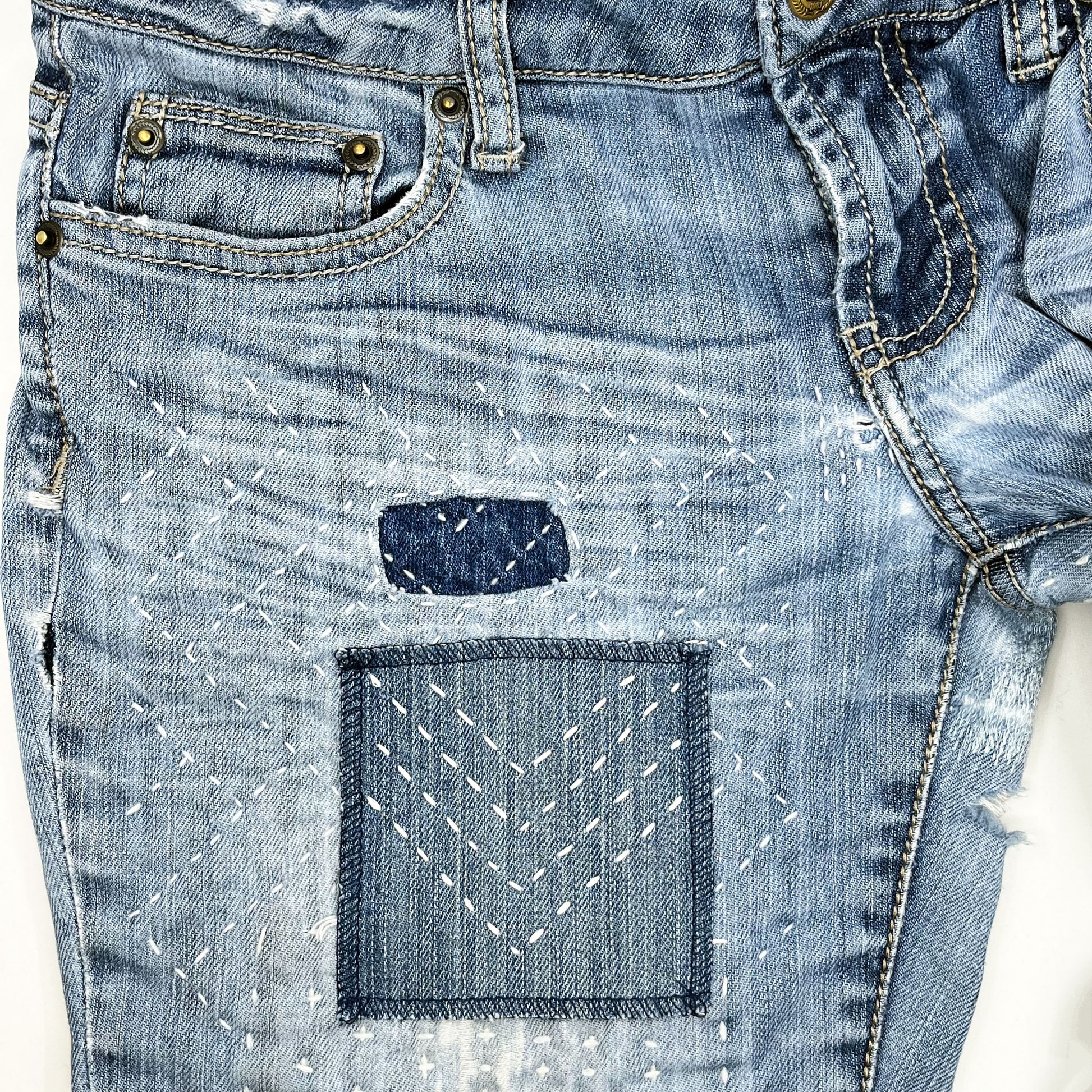 a square patch made out of denim, handstitched with rows of ivory running stitches in a chevron pattern, with overlocked edges, on a pair of jeans with other visible mending