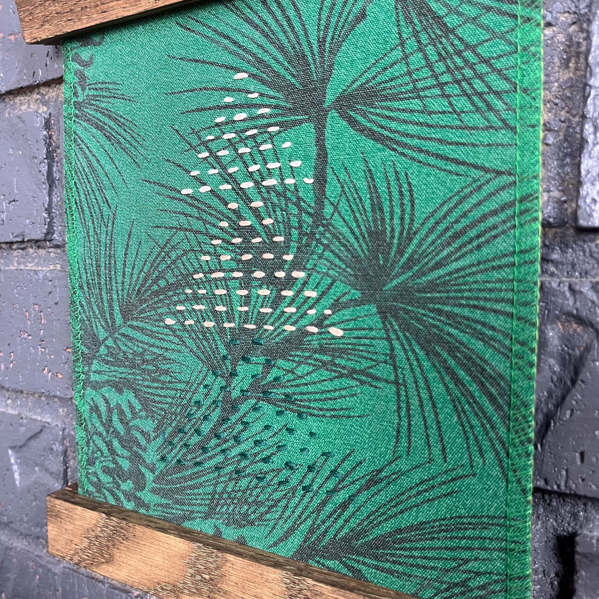 a fabric wall hanging in a magnetic wood frame, made from bright green fabric with pine branches printed on it, and stitched over with Christmas trees made from three triangles, each triangle made from running stitches in green peach and ivory