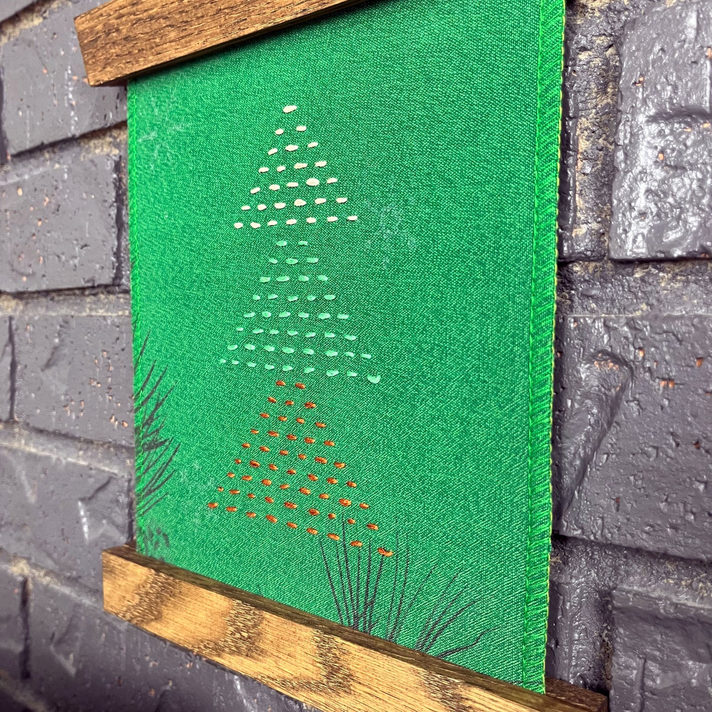 a fabric wall hanging in a magnetic wood frame, made from bright green fabric with pine branches printed on it, and stitched over with Christmas trees made from three triangles, each triangle made from running stitches in red green and peach