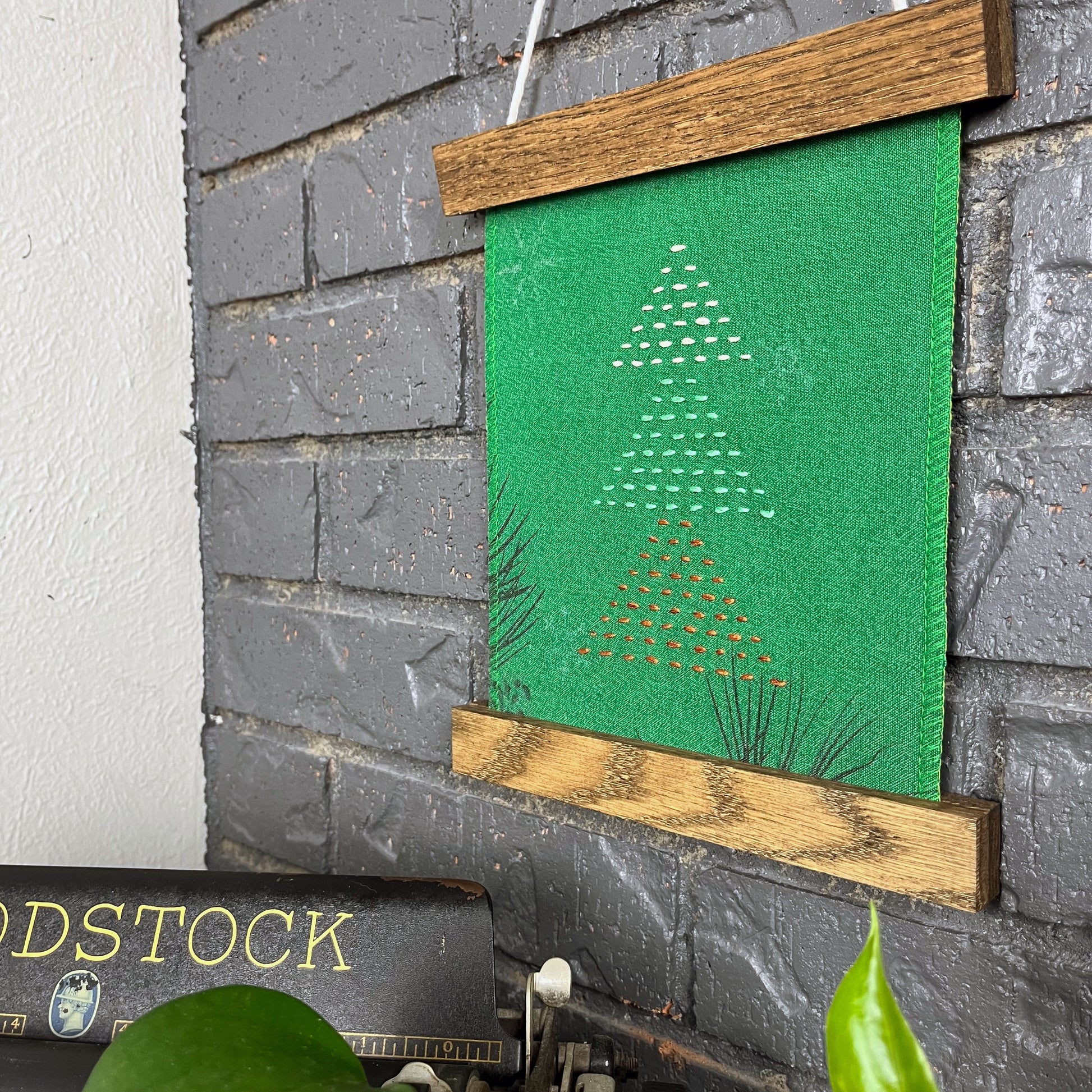 a fabric wall hanging in a magnetic wood frame, made from bright green fabric with pine branches printed on it, and stitched over with Christmas trees made from three triangles, each triangle made from running stitches in red green and peach