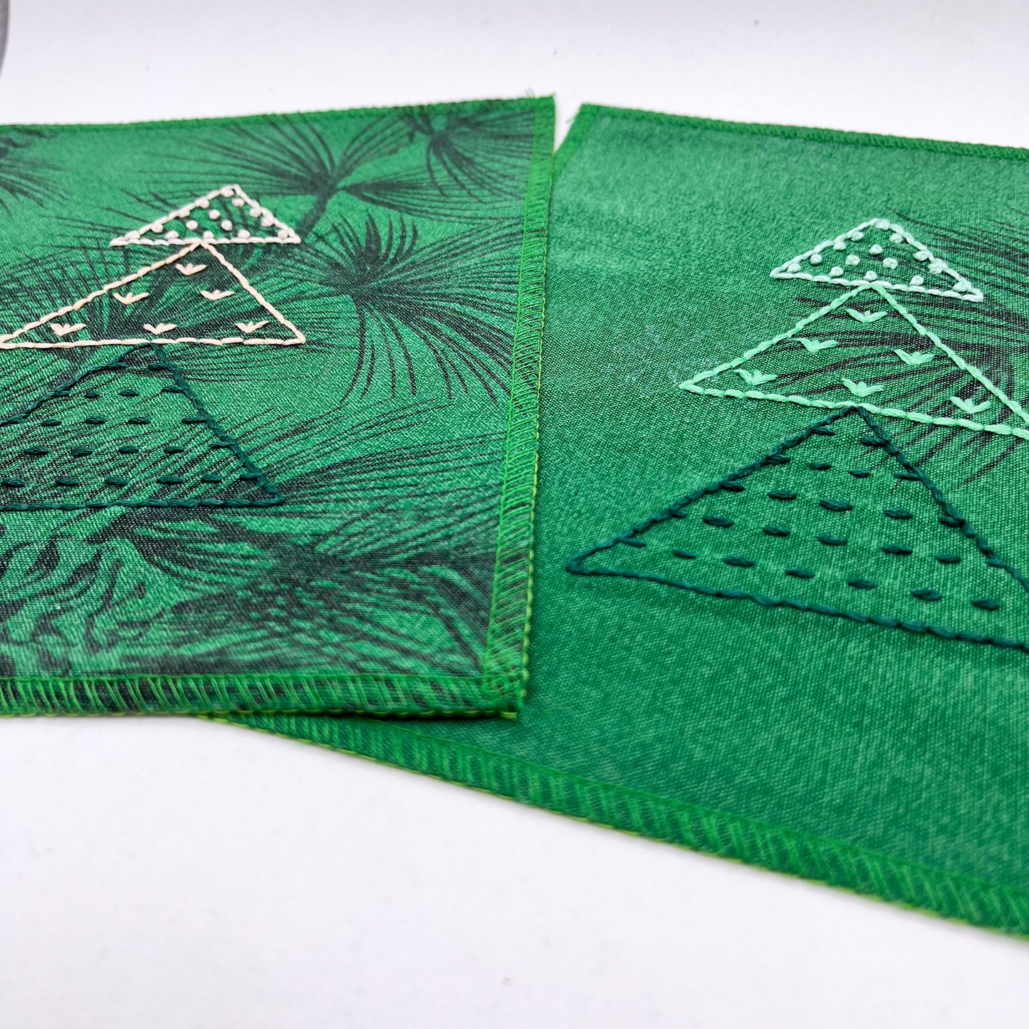 two fabric wall hangings made from bright green fabric with pine branches printed on them, and stitched over Christmas trees made from three triangles, each triangle filled with different types of stitches in different colors