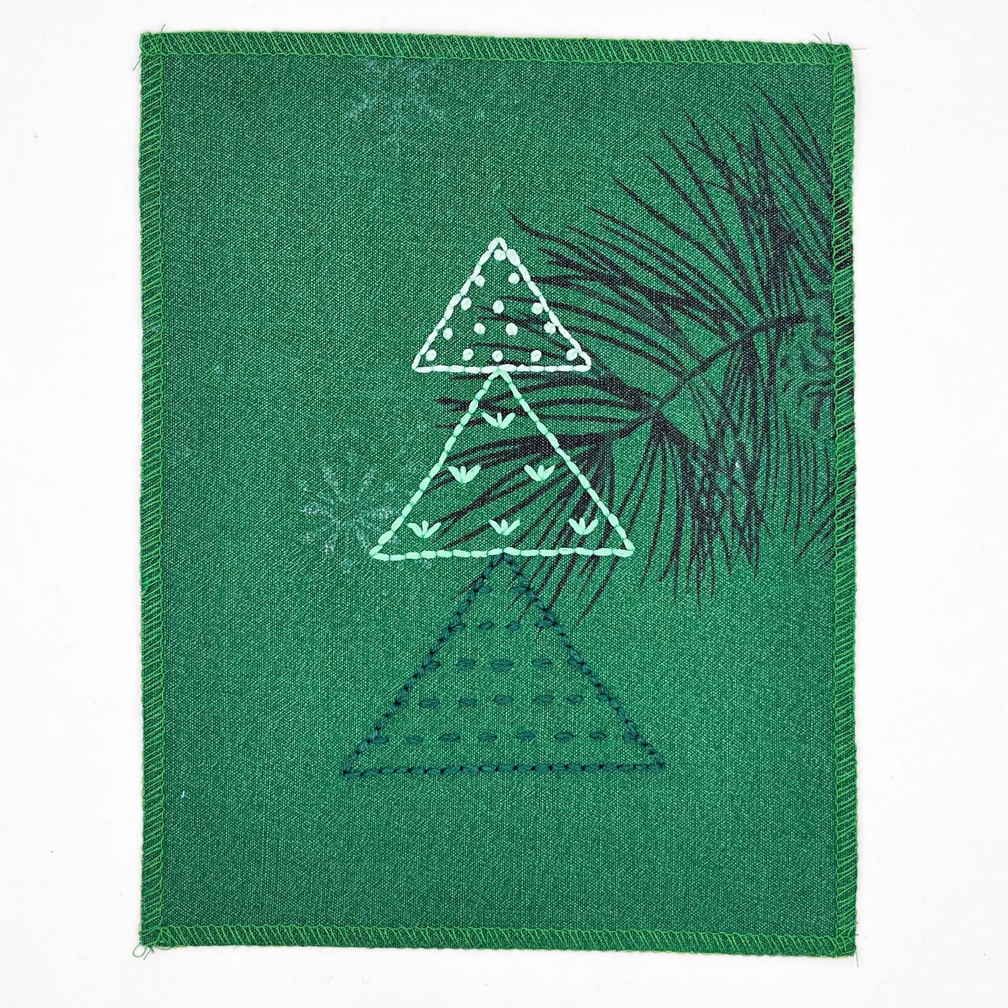 a fabric wall hanging made from bright green fabric with pine branches printed on it, and stitched over with Christmas trees made from three triangles, each triangle filled with different types of stitches in shades of green