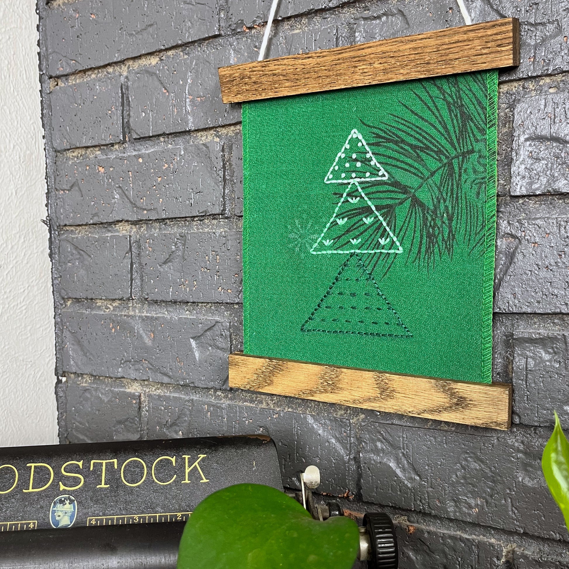 a fabric wall hanging in a magnetic wood frame, made from bright green fabric with a pine branch printed on it, and stitched over with Christmas trees made from three triangles, each triangle filled with different types of stitches in shades of green