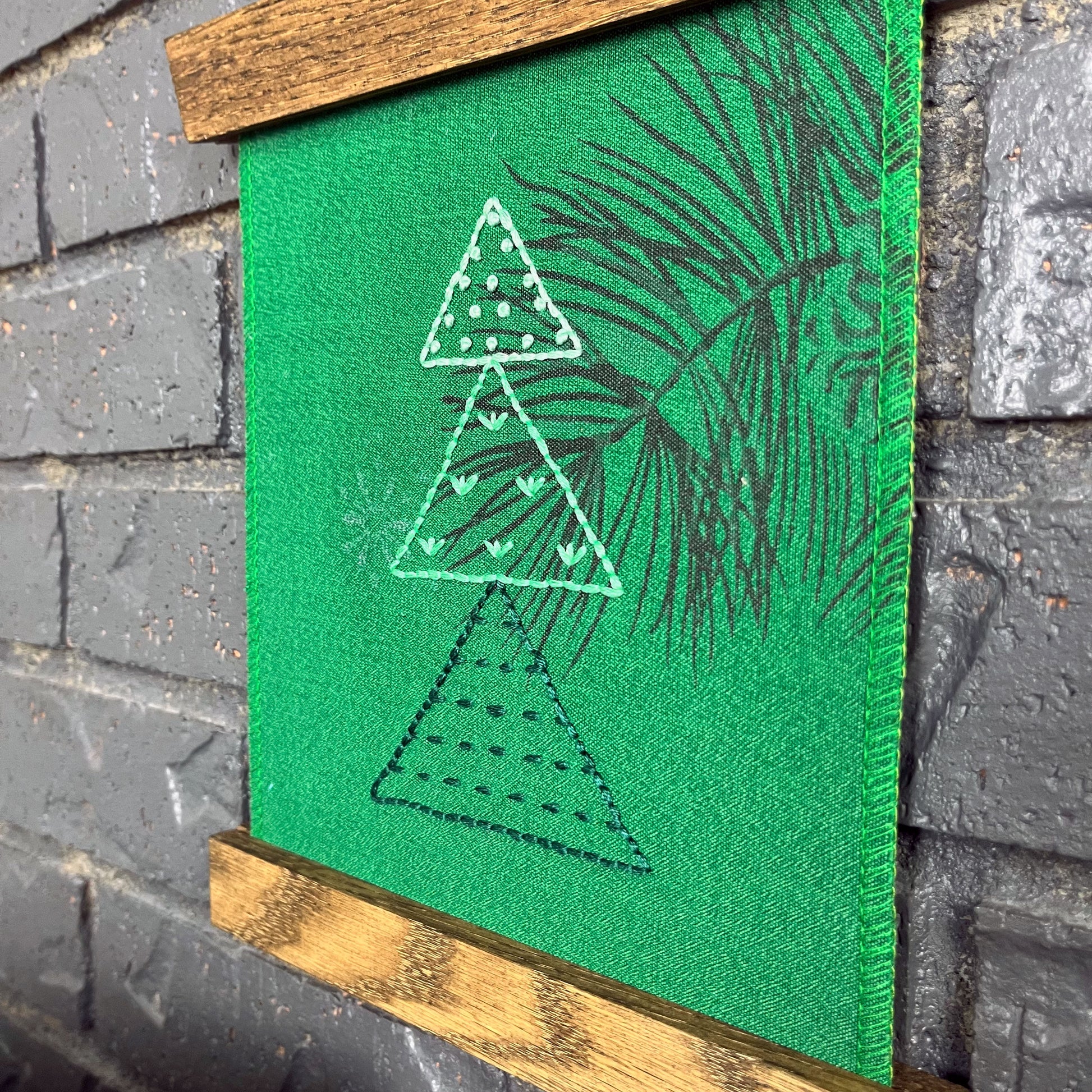 a fabric wall hanging in a magnetic wood frame, made from bright green fabric with a pine branch printed on it, and stitched over with Christmas trees made from three triangles, each triangle filled with different types of stitches in shades of green