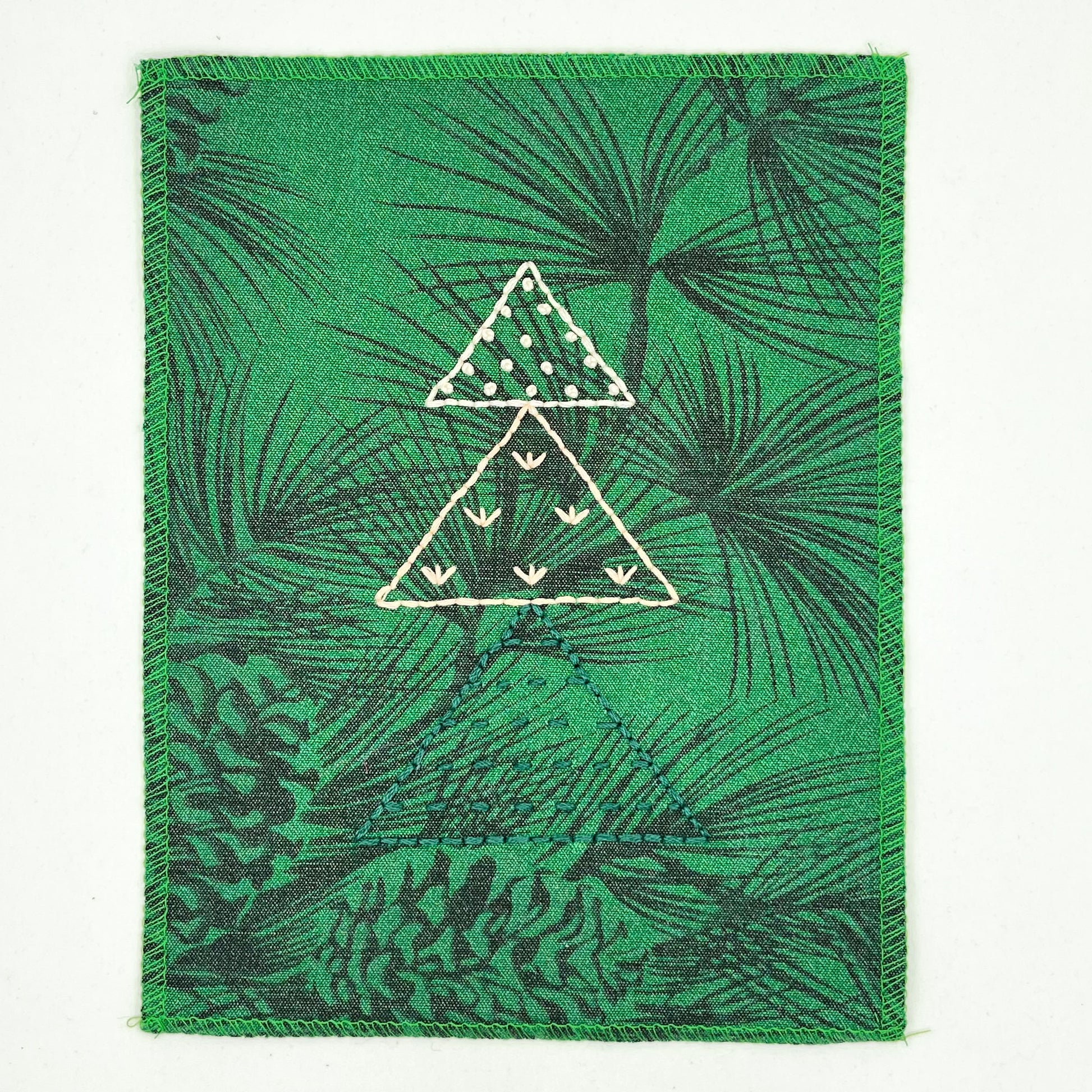a fabric wall hanging made from bright green fabric with pine branches printed on it, and stitched over with Christmas trees made from three triangles, each triangle filled with different types of stitches in green peach and ivory