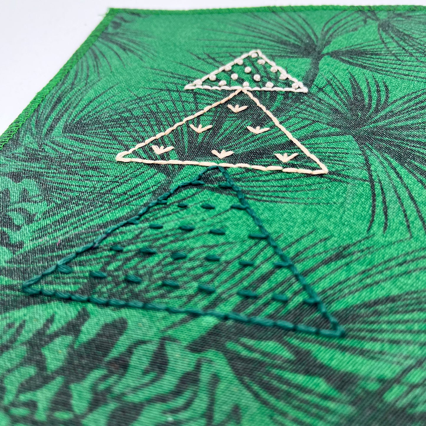 a close up view of a fabric wall hanging made from bright green fabric with pine branches printed on it, and stitched over with Christmas trees made from three triangles, each triangle filled with different types of stitches in green peach and ivory