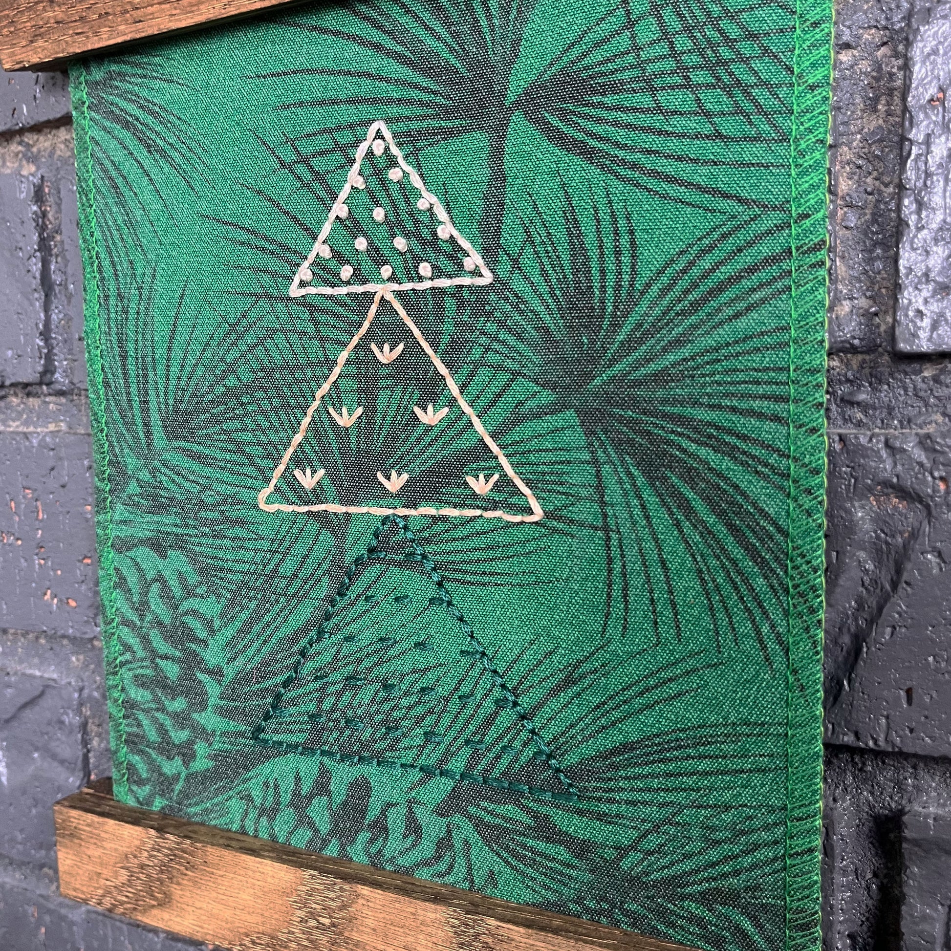 a fabric wall hanging in a magnetic wood frame, made from bright green fabric with pine branches printed on it, and stitched over with Christmas trees made from three triangles, each triangle filled with different stitches in green peach and ivory