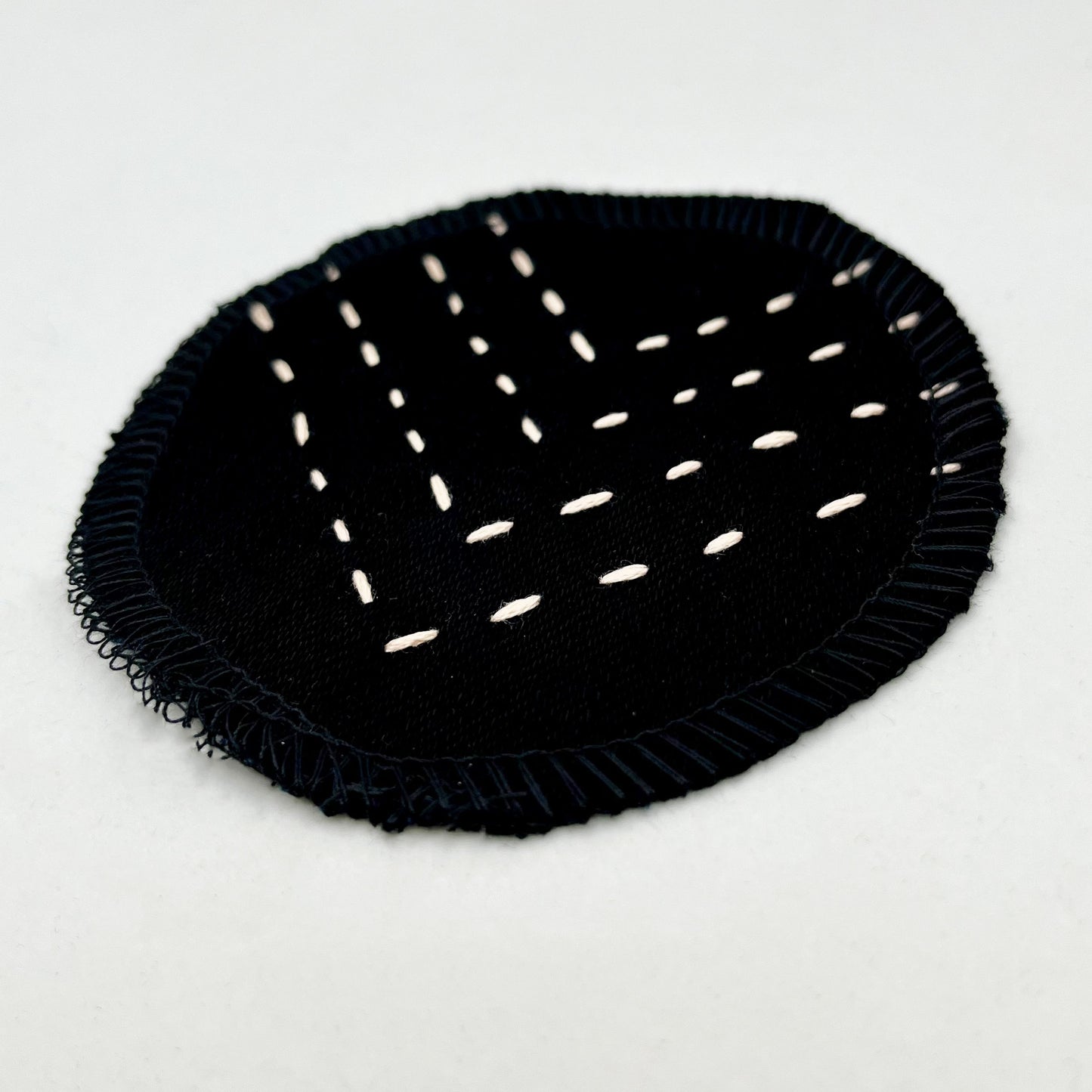 close up angled view of a circle shaped patch made out of black denim, with overlocked edges, with rows of peach running stitches in a chevron pattern, on a white background