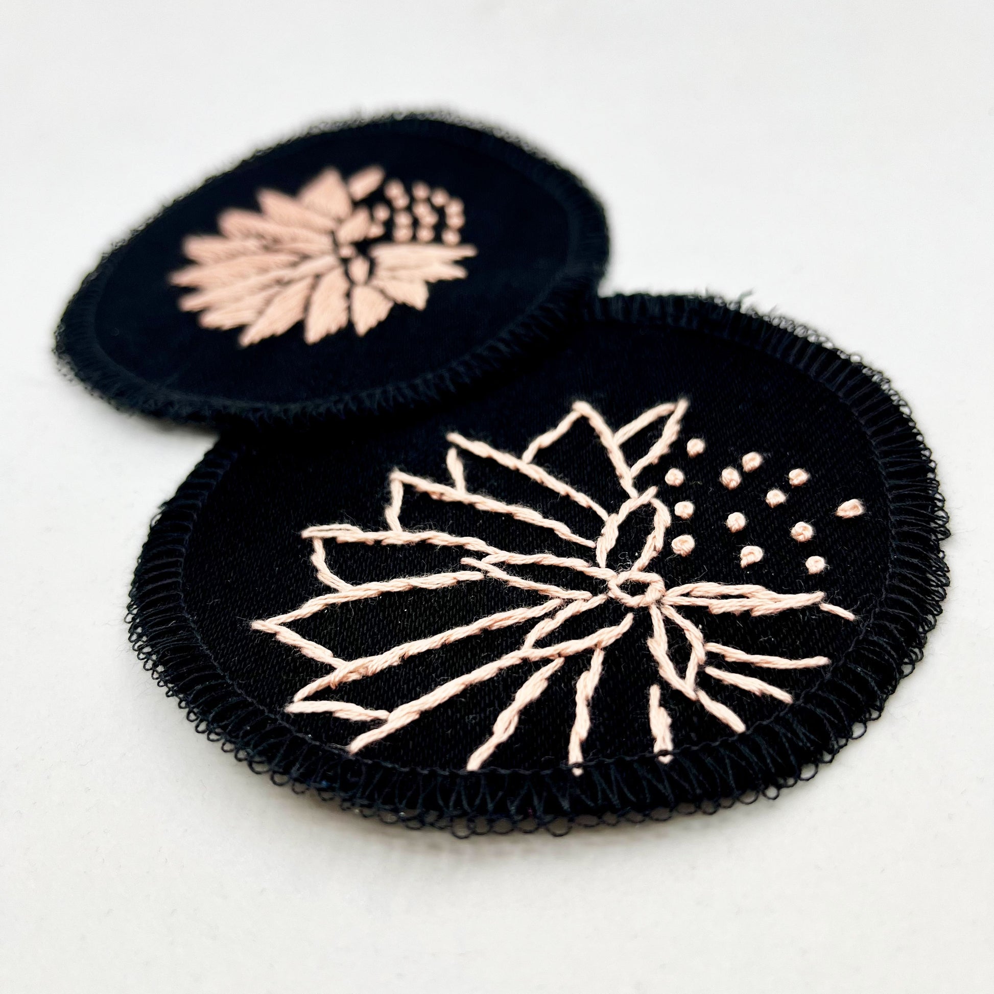 two circle shaped patches made out of black twill weave fabric, hand embroidered with abstract dandelions in peach colored floss, one with solid filled petals, the other with outlines of petals, edges overlocked in black, on a white background