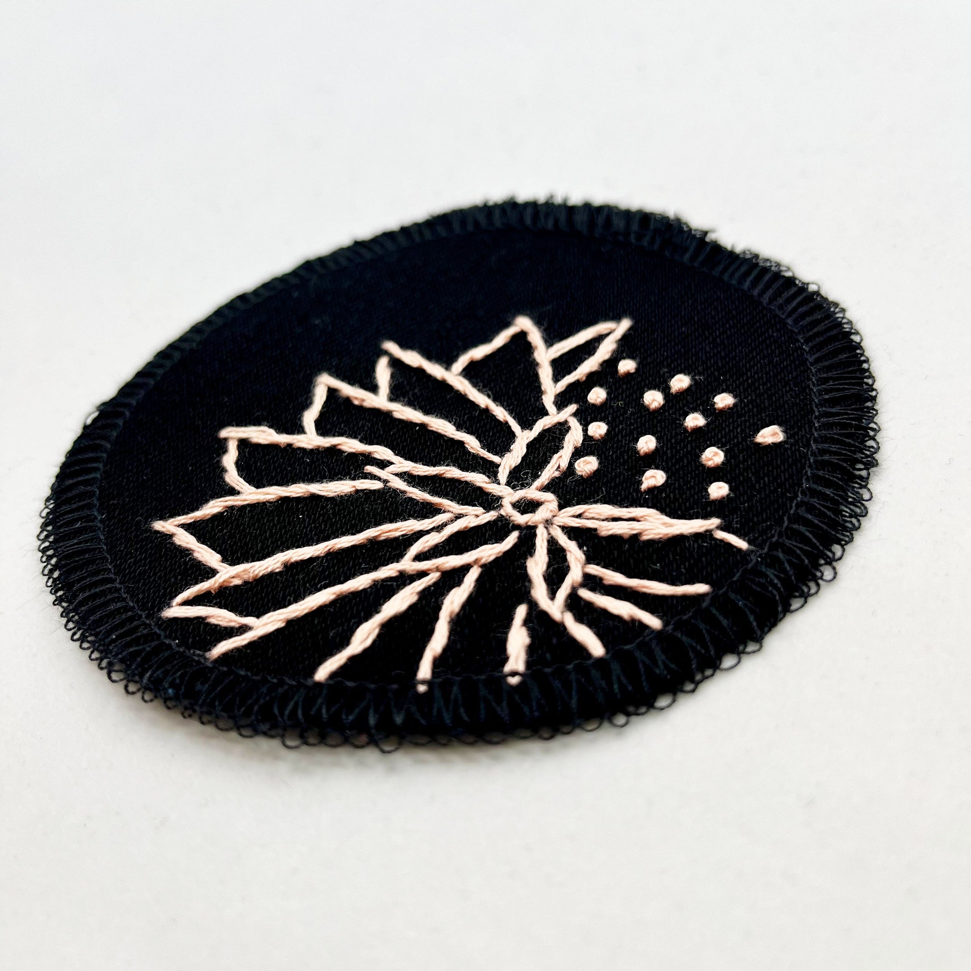 a circle shaped patch made out of black twill weave fabric, hand embroidered with an abstract dandelion in peach colored floss, outlines of petals, edges overlocked in black, on a white background