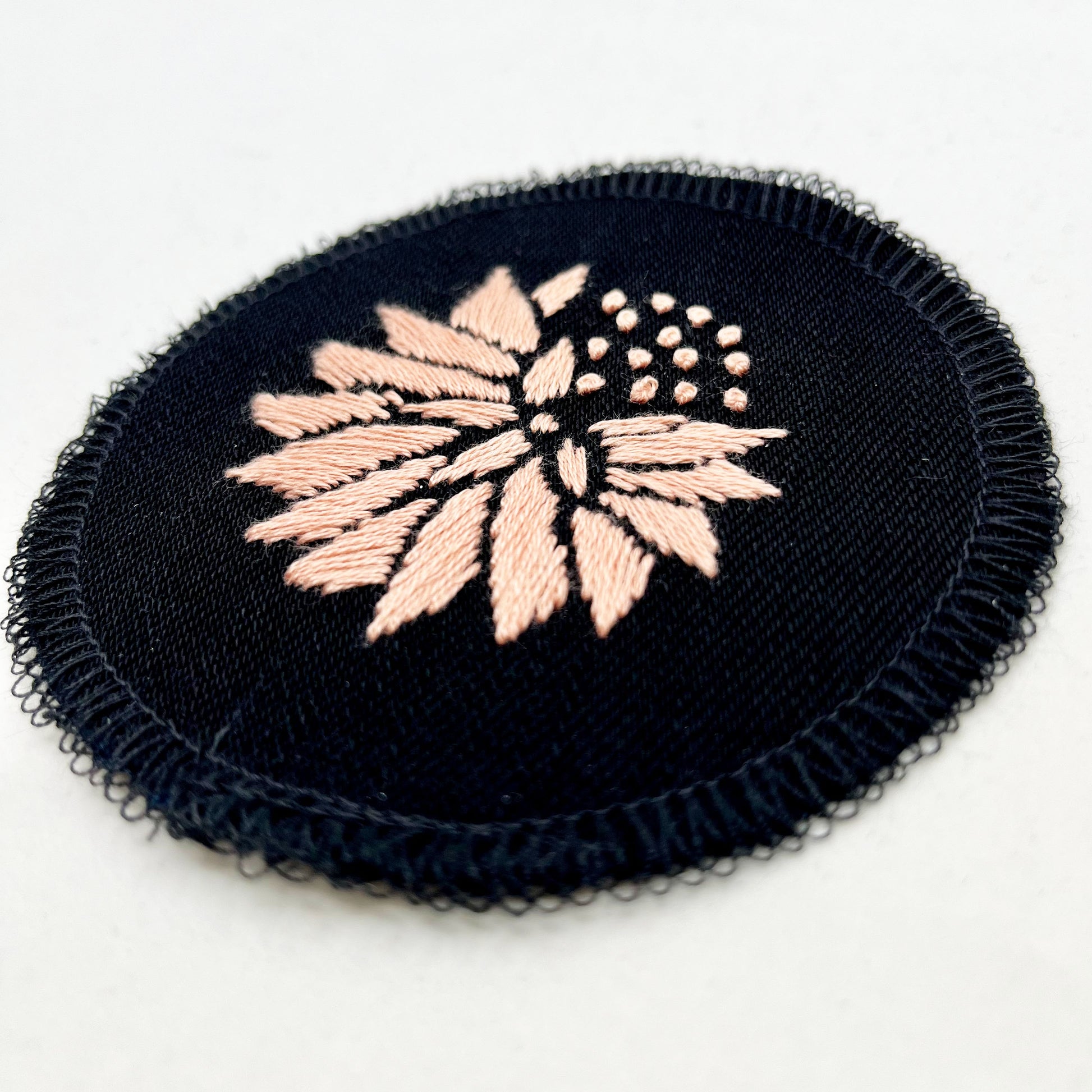 a circle shaped patch made out of black twill weave fabric, hand embroidered with an abstract dandelion in peach colored floss, with solid filled petals, edges overlocked in black, on a white background