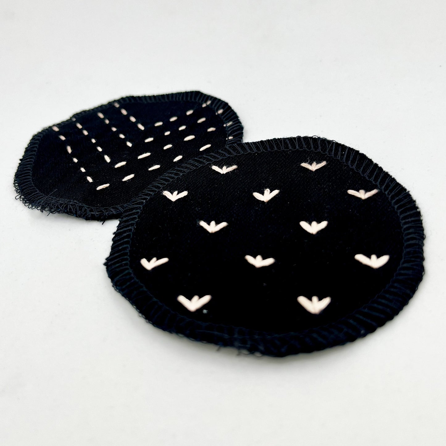 close up angled view of two circle shaped patches made out of black denim, with overlocked edges, one with stitches in peach that look like sprouts or birds feet, the other with rows of running stitches in a chevron pattern