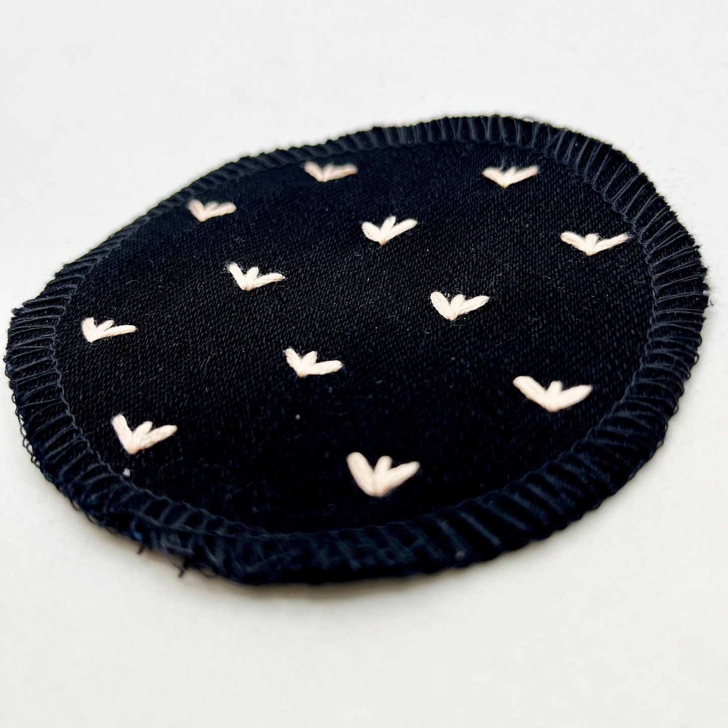 a close up angled view of a circle shaped patch made out of black denim, with overlocked edges, with stitches in peach that look like sprouts or birds feet, on a white background