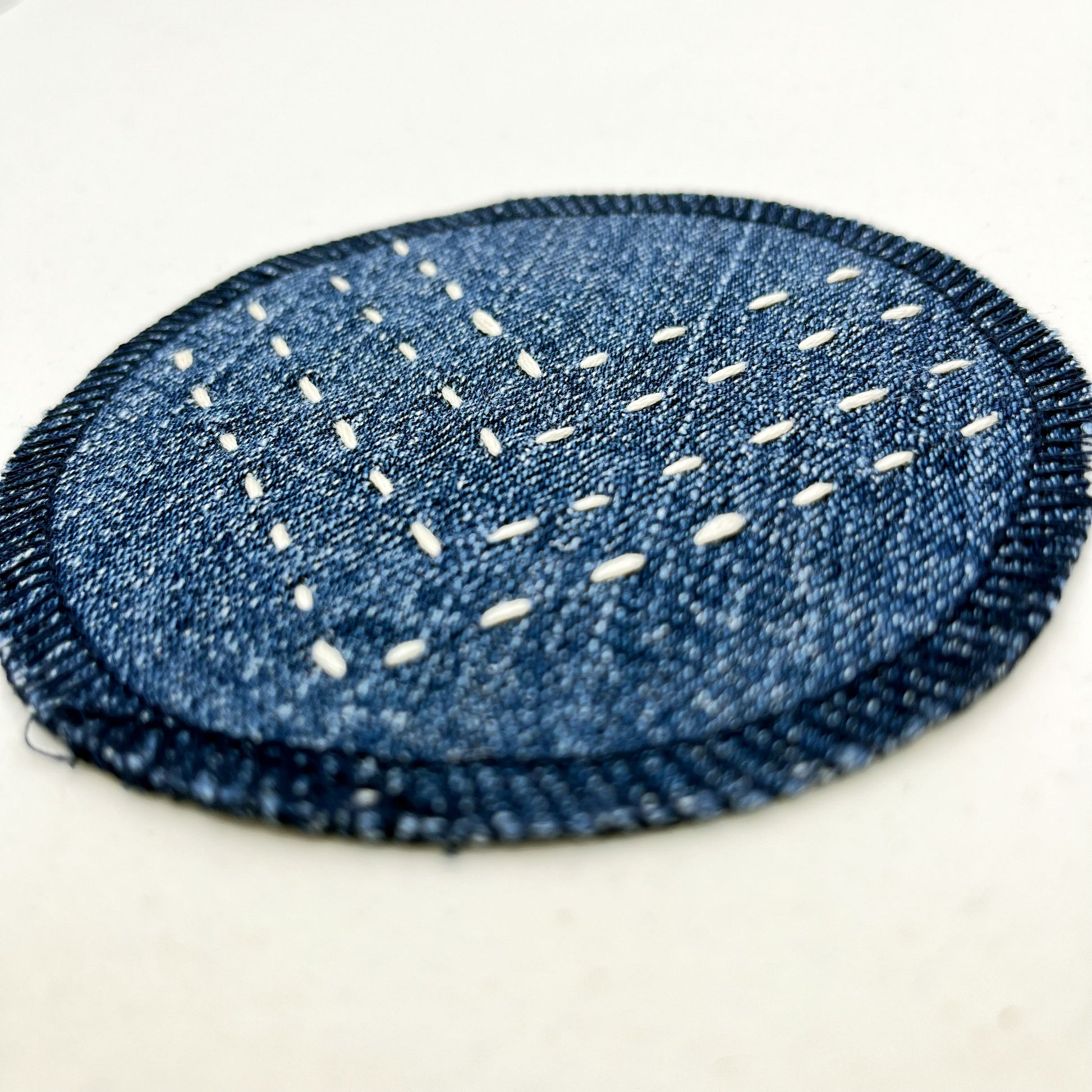 a close up angled view of a circle shaped patch made out of denim, with overlocked edges, with rows of ivory running stitches in a chevron pattern, on a white background