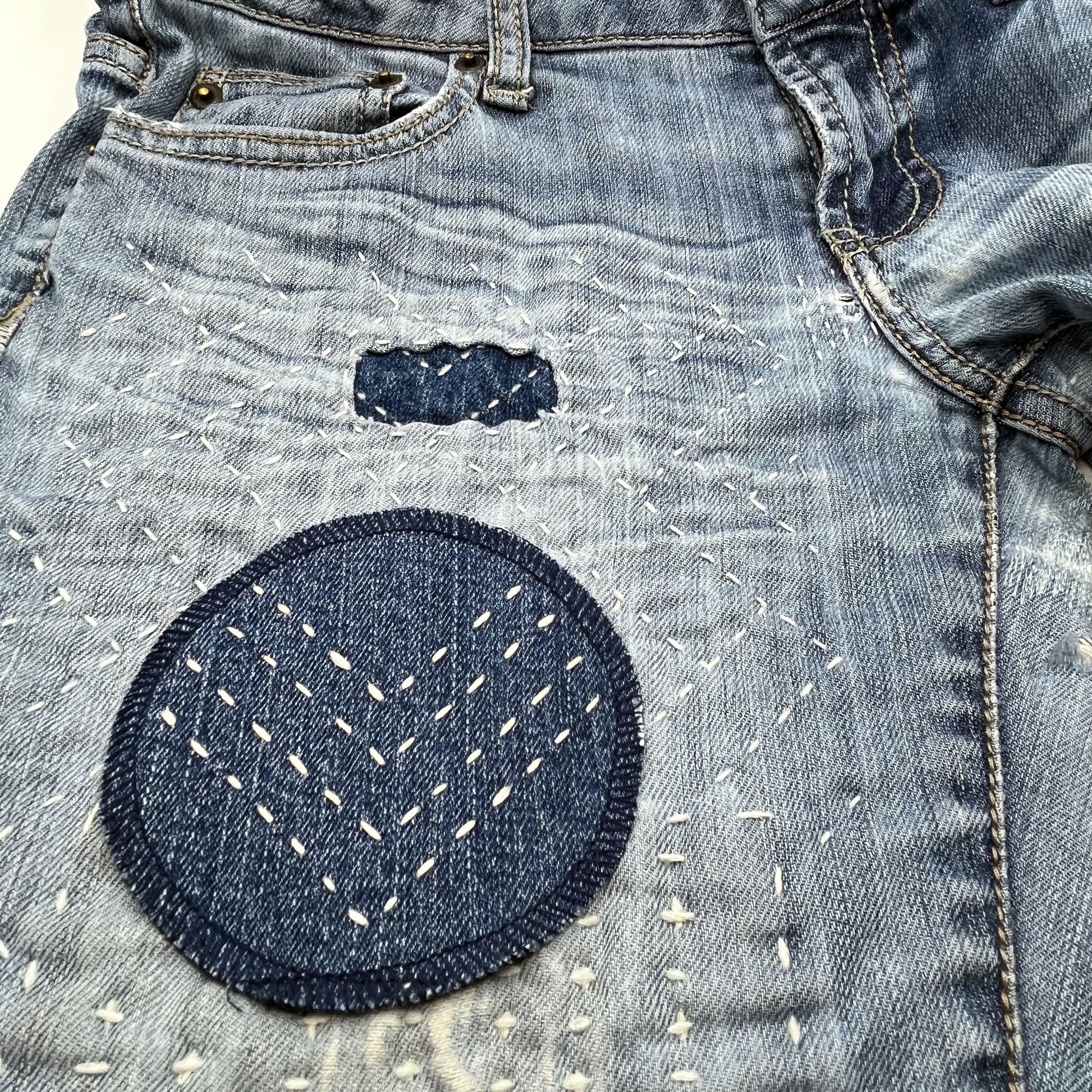 close up angled view of a circle shaped patch made out of denim, with overlocked edges, with rows of running stitches in a chevron pattern, on a pair of jeans with other visible mending and sashiko stitching