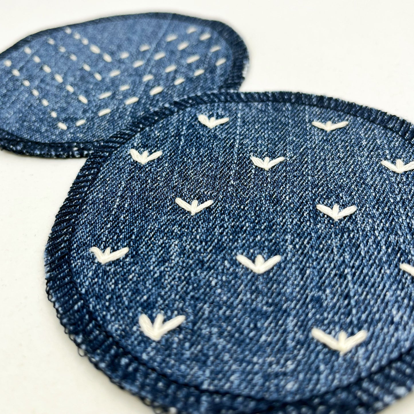 close up angled view of two circle shaped patches made out of denim, with overlocked edges, one with stitches in ivory that look like sprouts or birds feet, the other with rows of running stitches in a chevron pattern