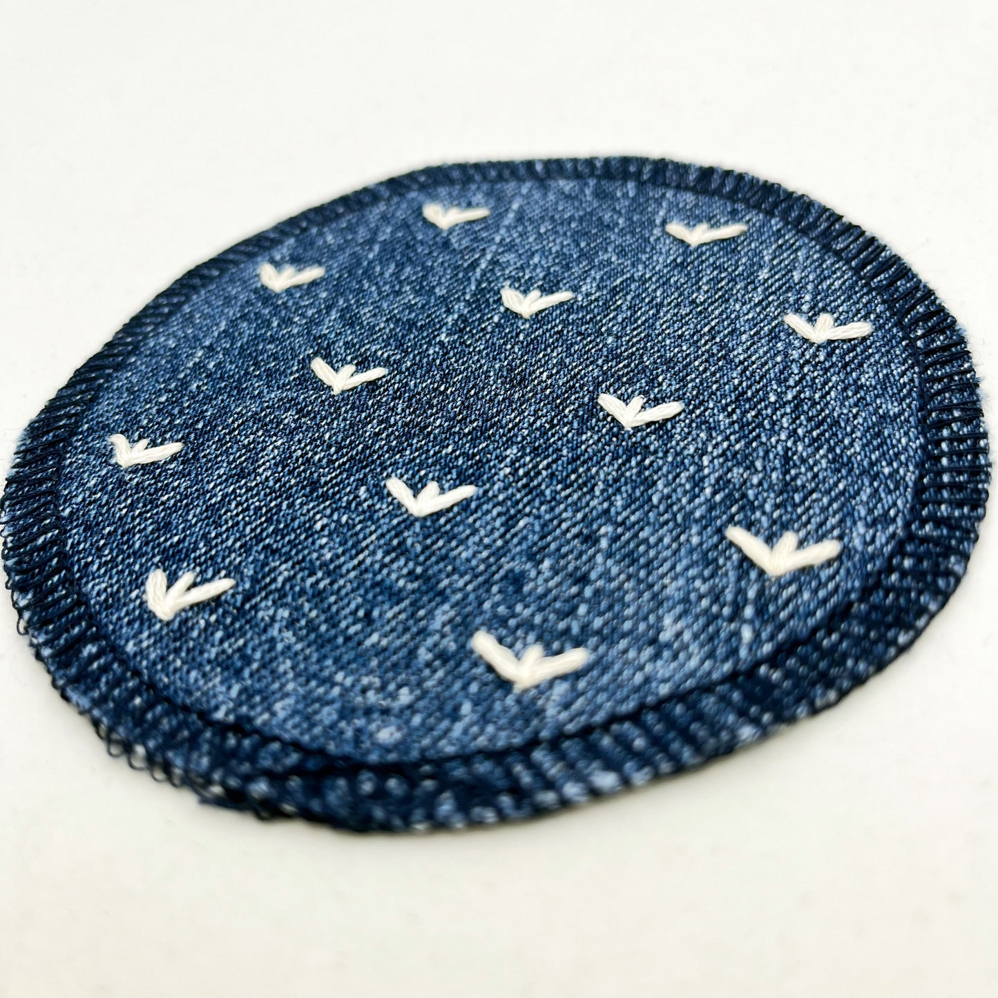 a close up angled view of a circle shaped patch made out of denim, with overlocked edges, with stitches in ivory that look like sprouts or birds feet, on a white background