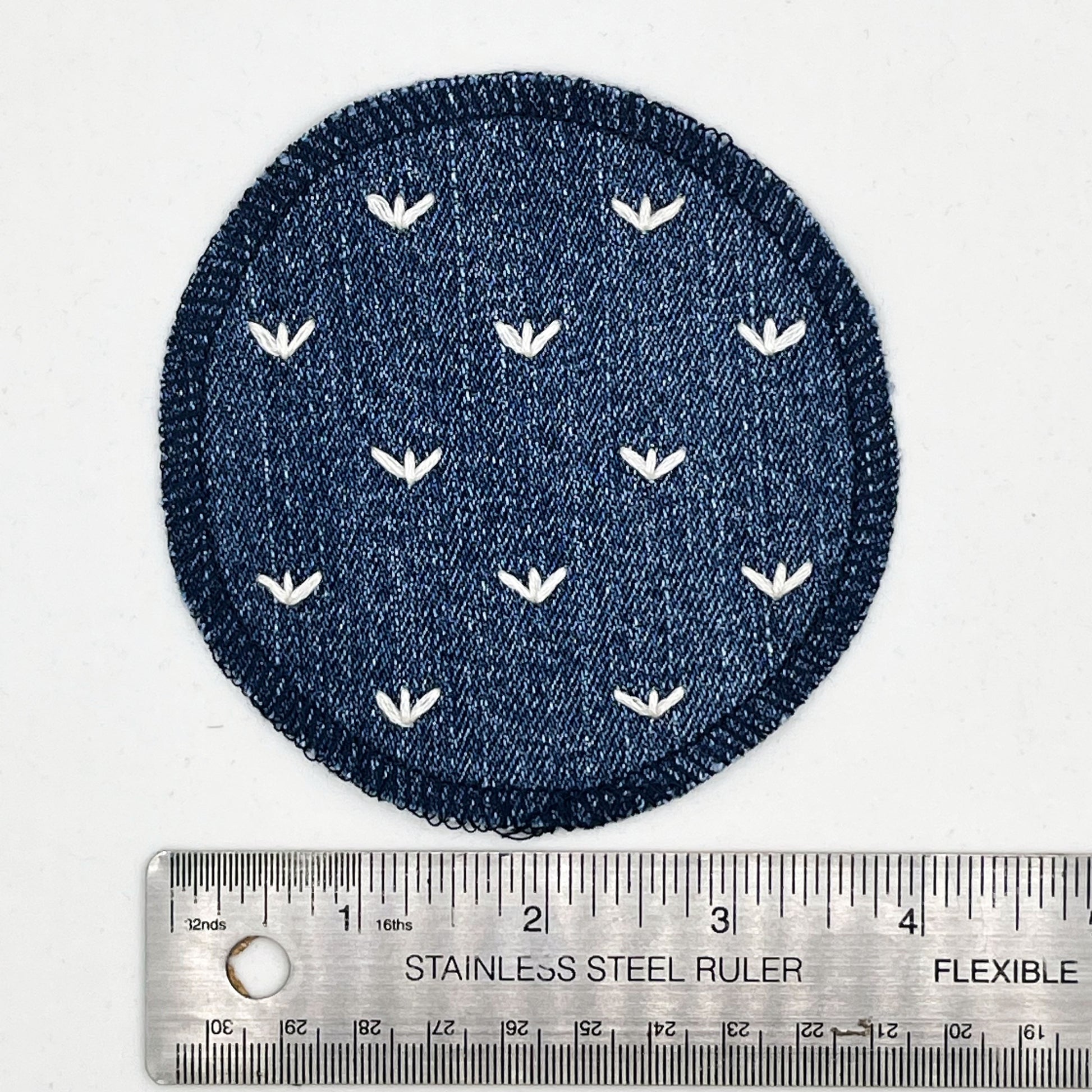 a circle shaped patch made out of denim, with overlocked edges, with stitches in ivory that look like sprouts or birds feet, next to a metal ruler to show a diameter or 4 inches, on a white background