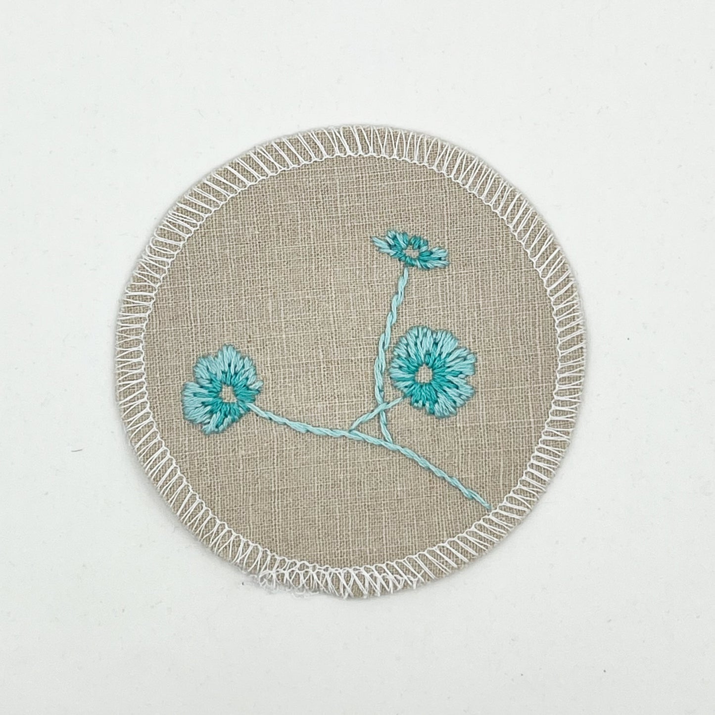 a circle shaped patch made from a natural color linen fabric, hand embroidered with 3 poppies in shades of robin's egg blues, edges overlocked with white thread, on a white background