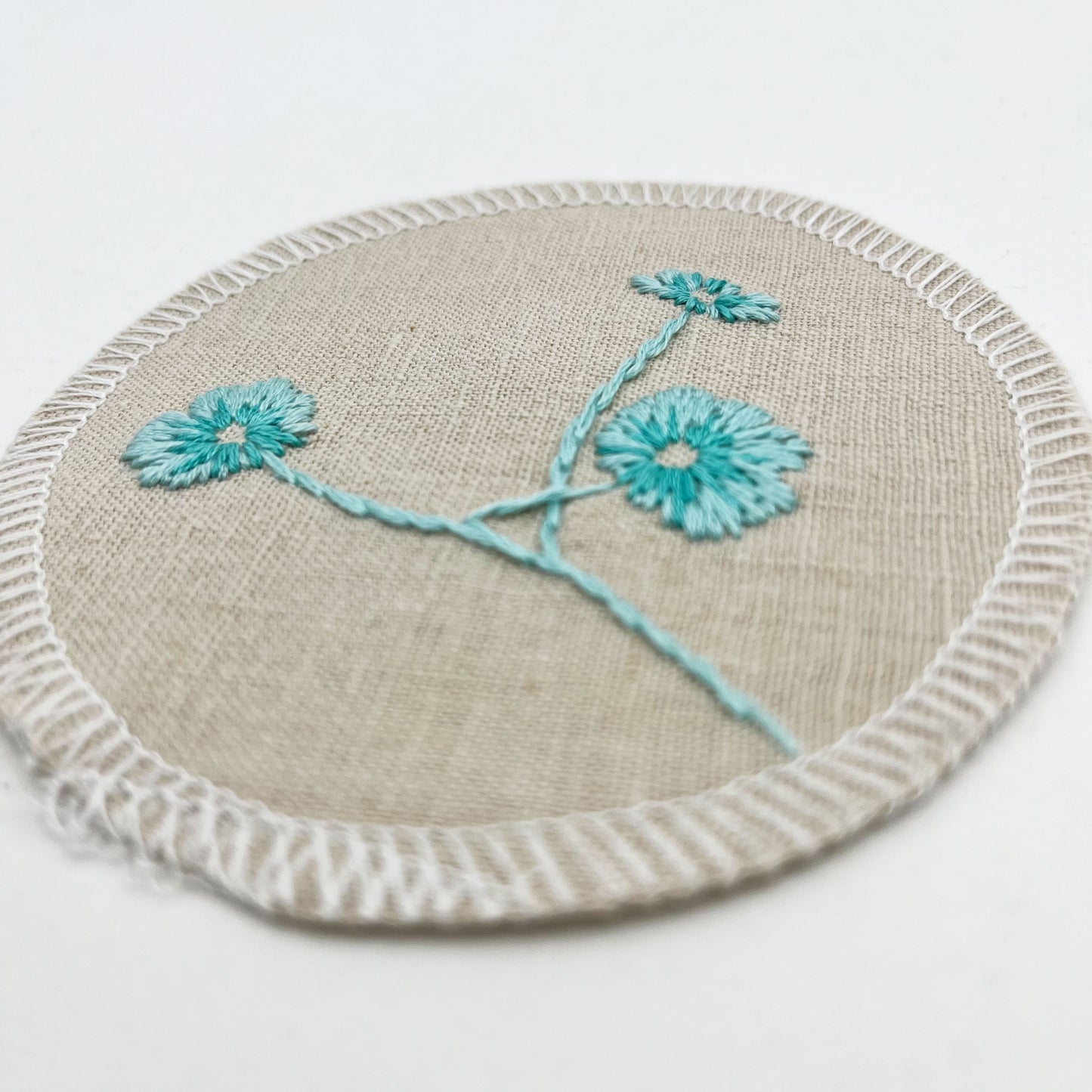 close up angled view of a circle shaped patch, made from a natural color linen fabric, hand embroidered with 3 poppies in shades of robin's egg blues, edges overlocked with white thread, on a white background