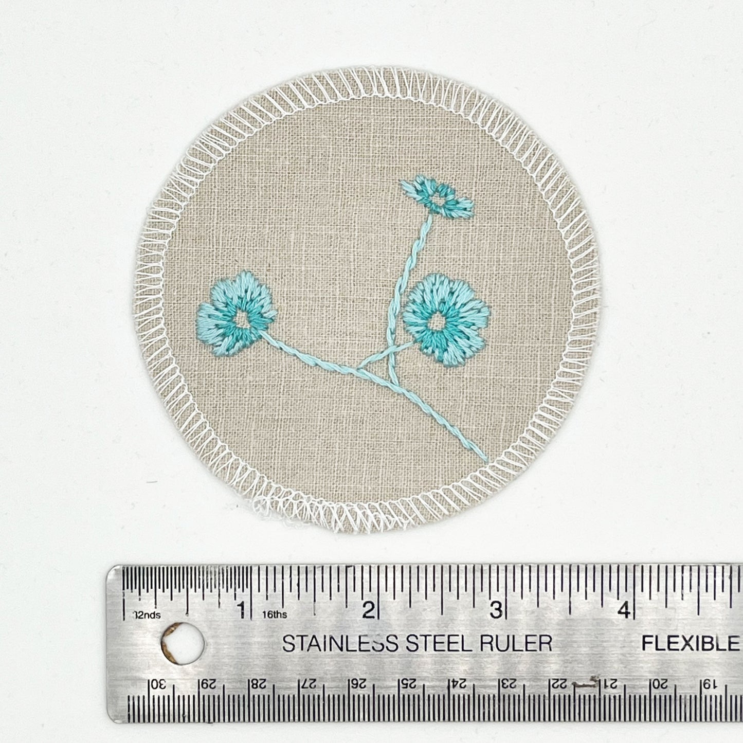 a patch made from a natural color cotton linen fabric placed next to a ruler, showing a diameter of 4 inches, hand embroidered with 3 poppies in shades of robin's egg blue, edges overlocked with white thread, on a white background