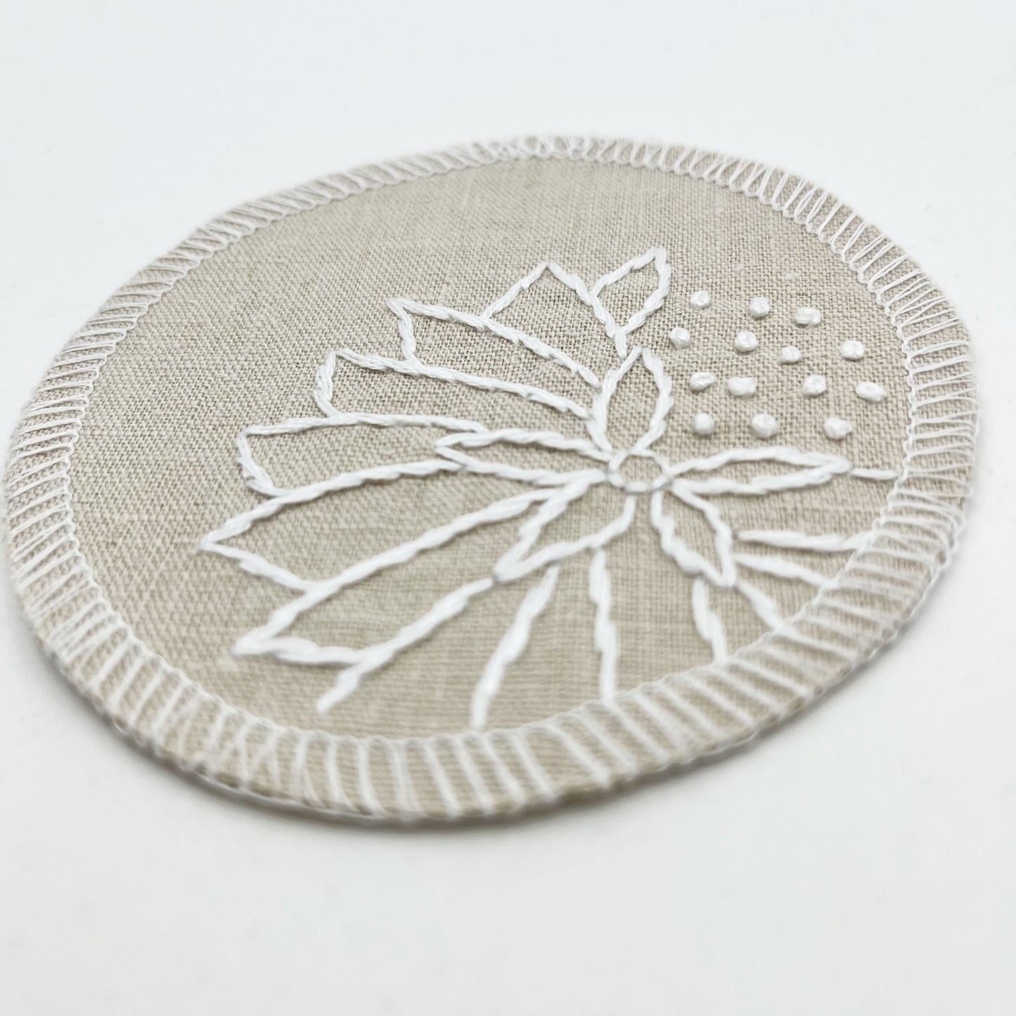 a close up angled view of a circle shaped patch made out of natural colored cotton linen fabric, hand embroidered with an abstract dandelion in white colored floss, outlines of petals, edges overlocked in white, on a white background
