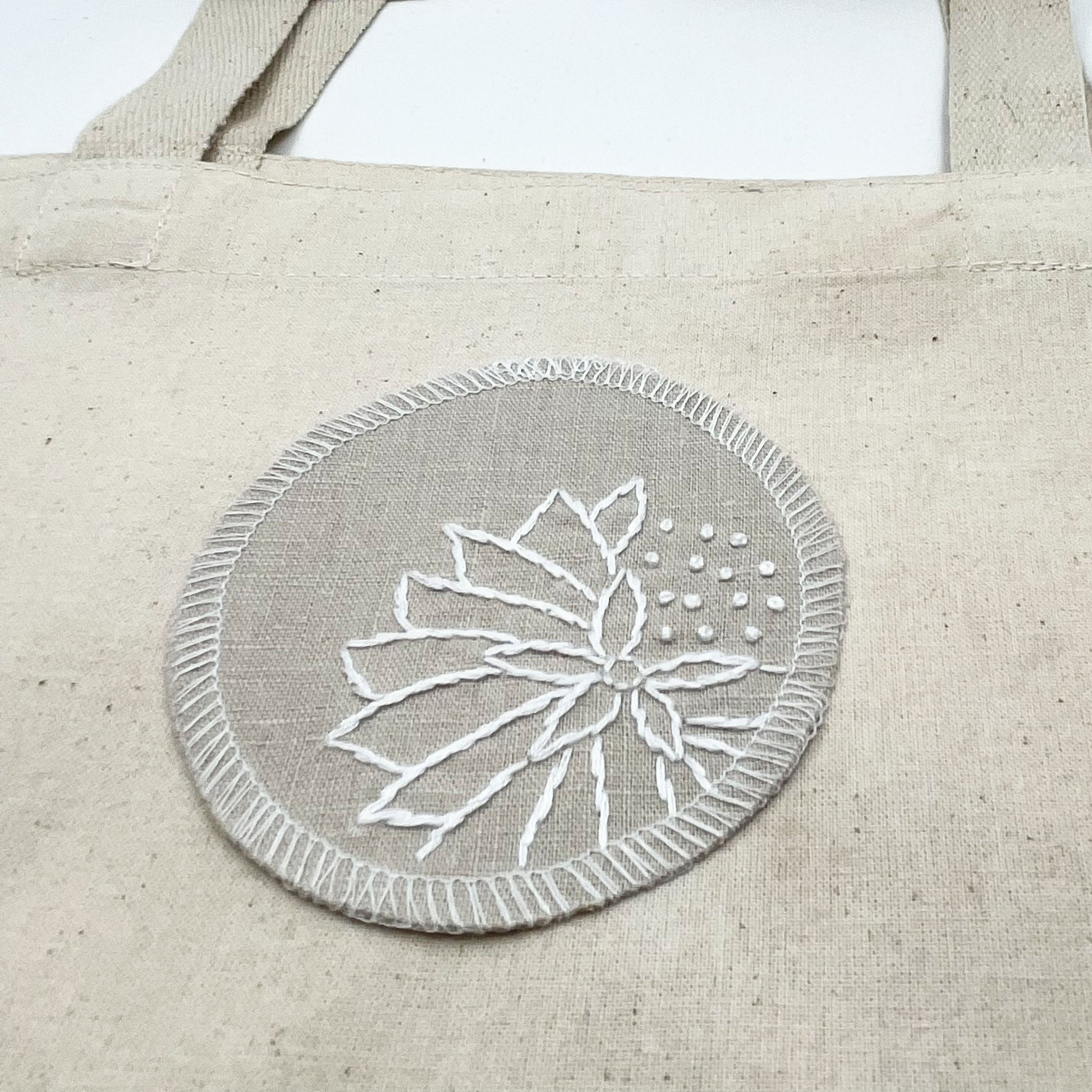 a circle shaped patch made out of natural colored cotton linen fabric, sitting on a canvas tote bag, hand embroidered with an abstract dandelion in white colored floss, outlines of petals, edges overlocked in white, on a white background