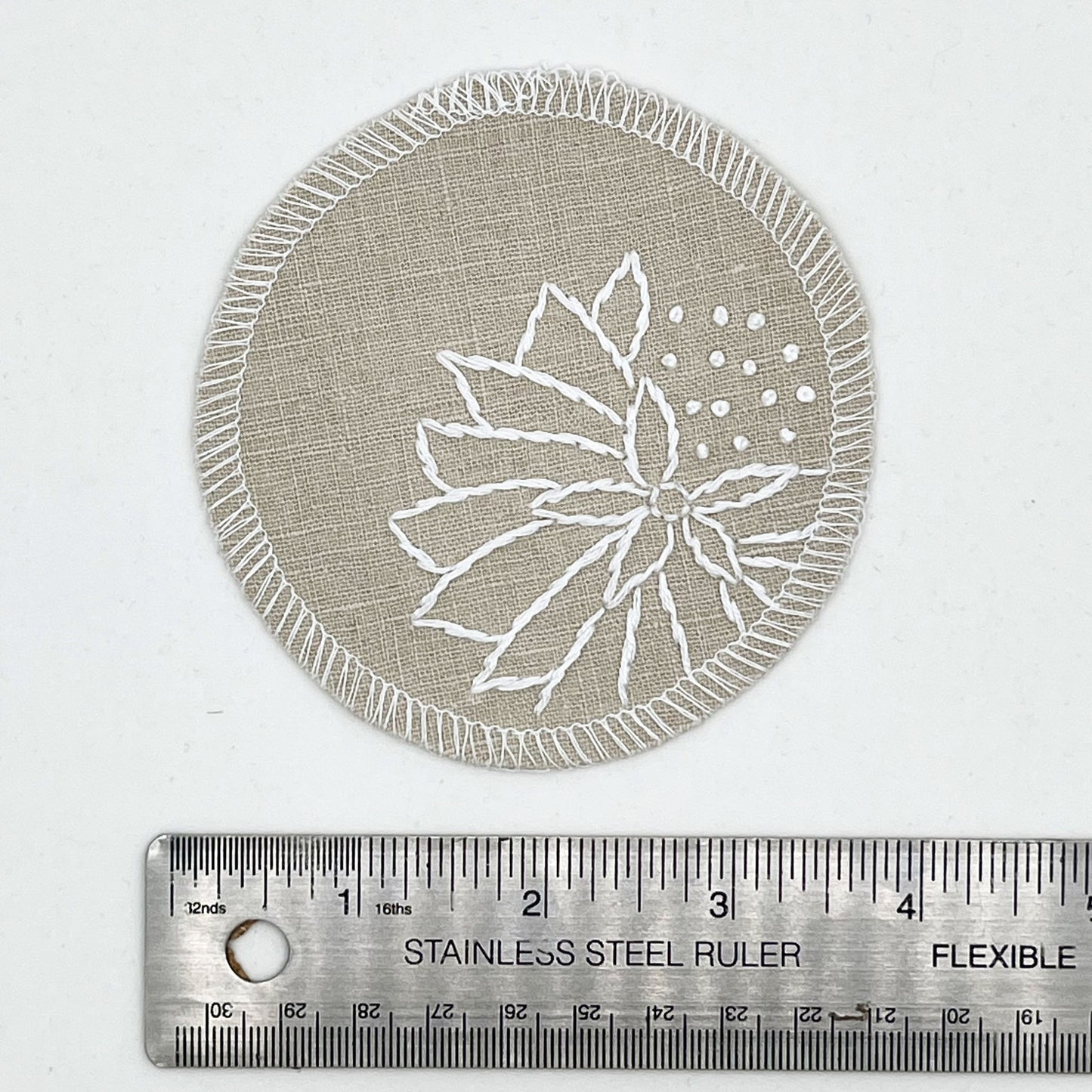 a circle shaped patch made out of natural colored cotton linen fabric, hand embroidered with an abstract dandelion in white colored floss, outlines of petals, edges overlocked in white, on a white background, placed next to a ruler to show a diameter of 4 inches