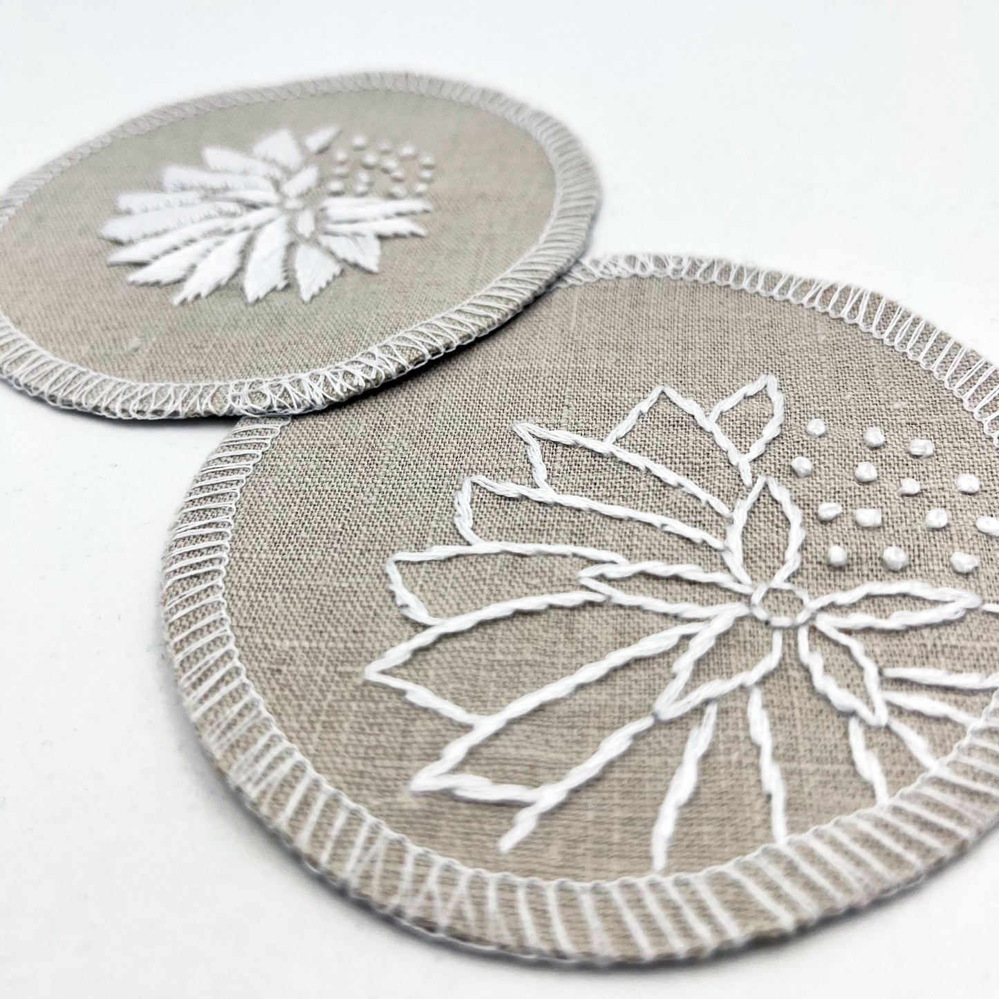 two circle shaped patches made out of natural colored cotton linen fabric, hand embroidered with abstract dandelions in white floss, one with solid filled petals, the other with outlines of petals, edges overlocked in white, on a white background