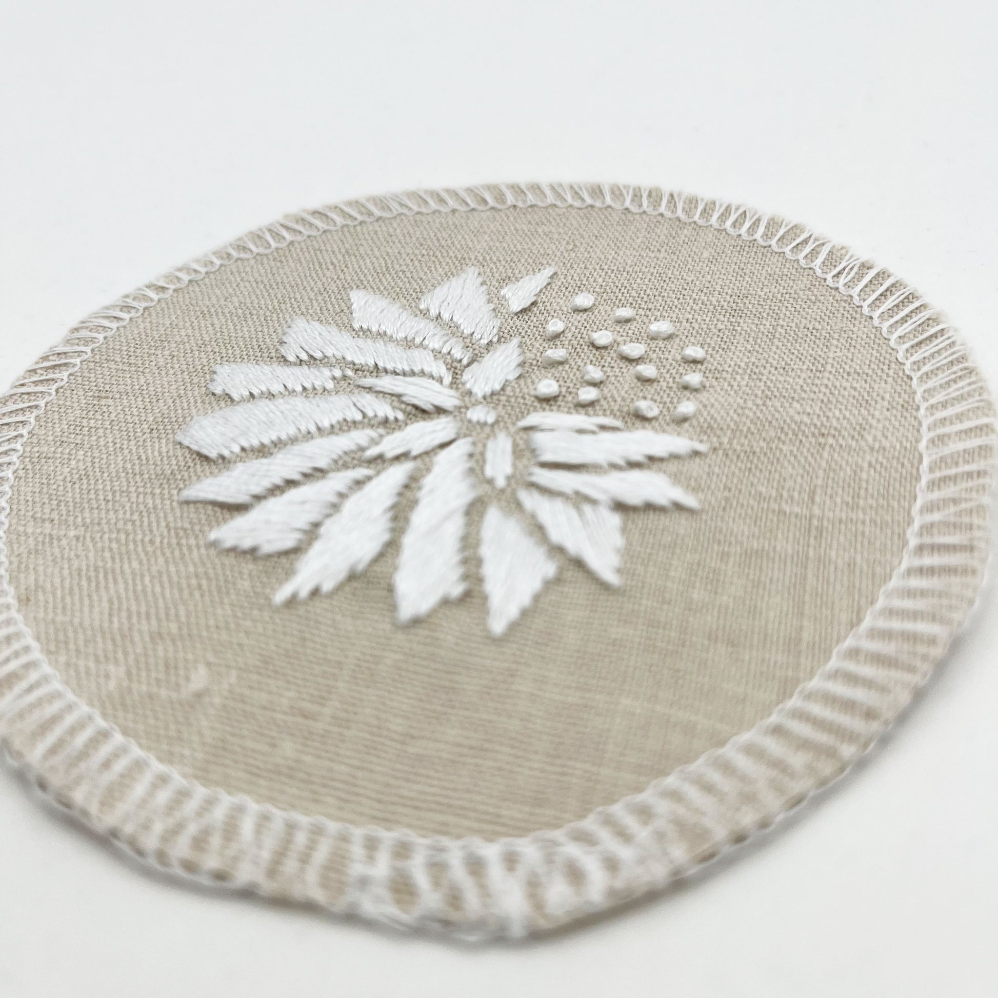 close up angled view of a circle shaped patch made out of natural colored cotton linen fabric, hand embroidered with an abstract dandelion in white colored floss, solid filled petals, edges overlocked in white, on a white background