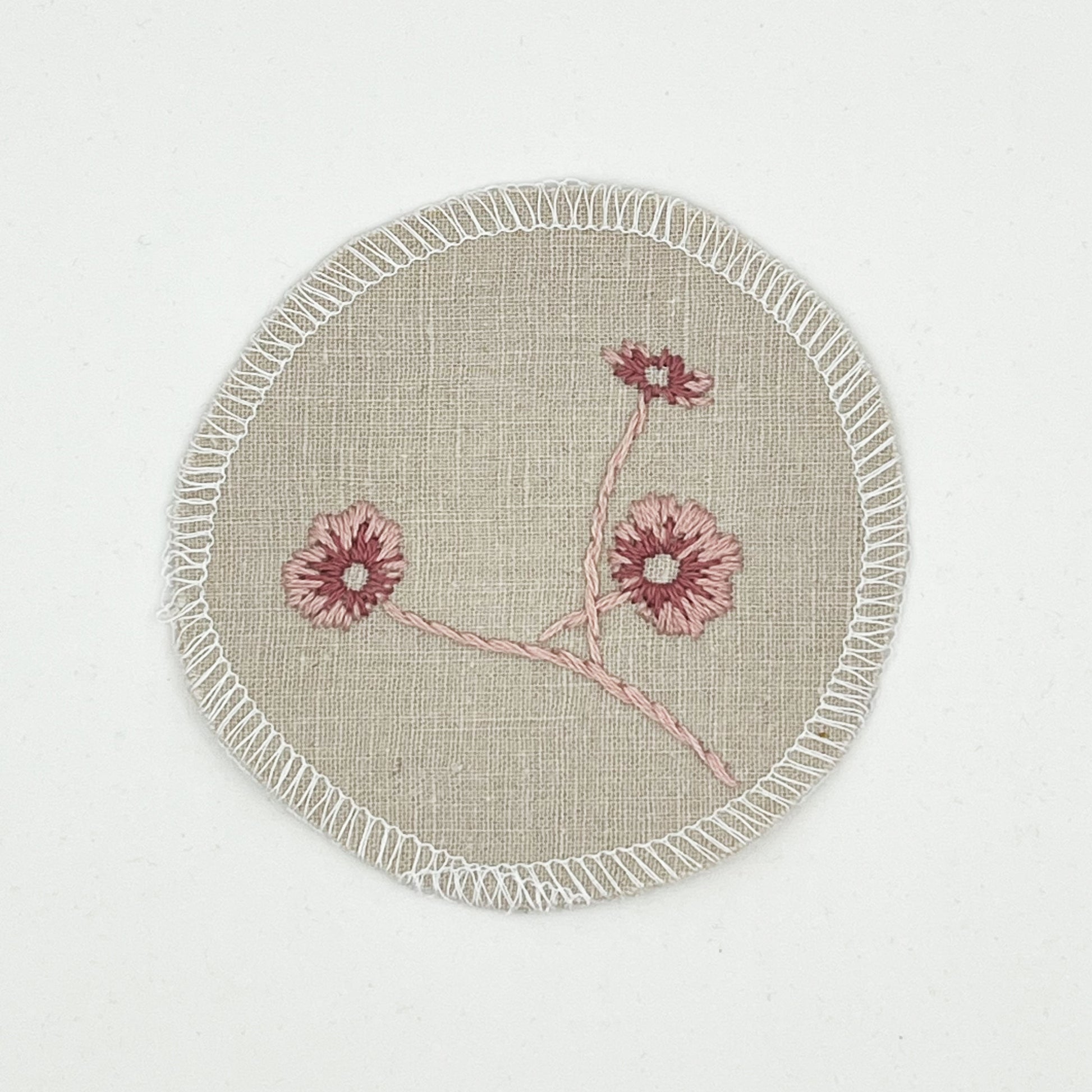 a circle shaped patch made from a natural color linen fabric, hand embroidered with 3 poppies in shades of pink, edges overlocked with white thread, on a white background