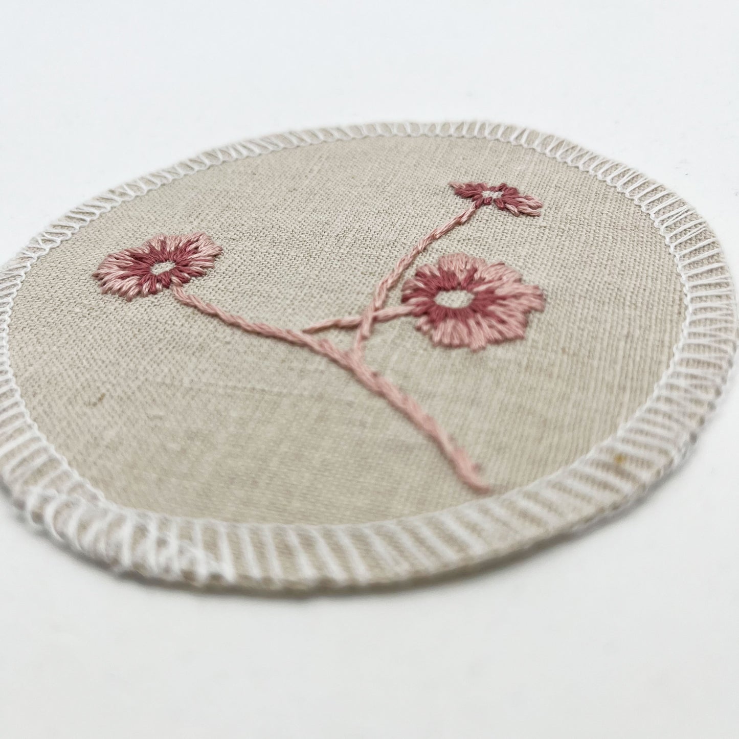 close up angled view of a circle shaped patch, made from a natural color linen fabric, hand embroidered with 3 poppies in shades of pink, edges overlocked with white thread, on a white background