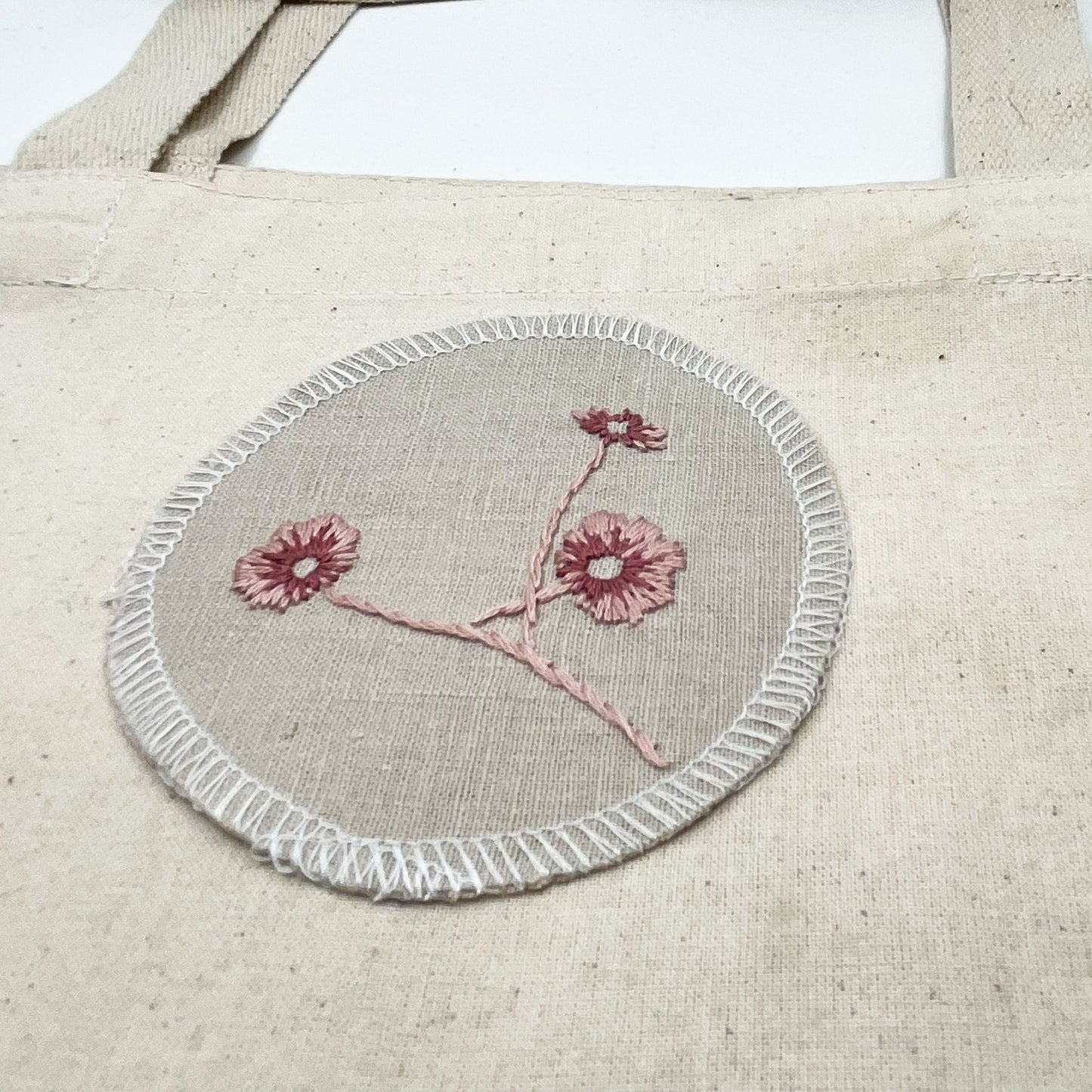 a patch made from a natural color linen fabric on a canvas tote bag, hand embroidered with 3 poppies in shades of pink, edges overlocked with white thread, on a white background