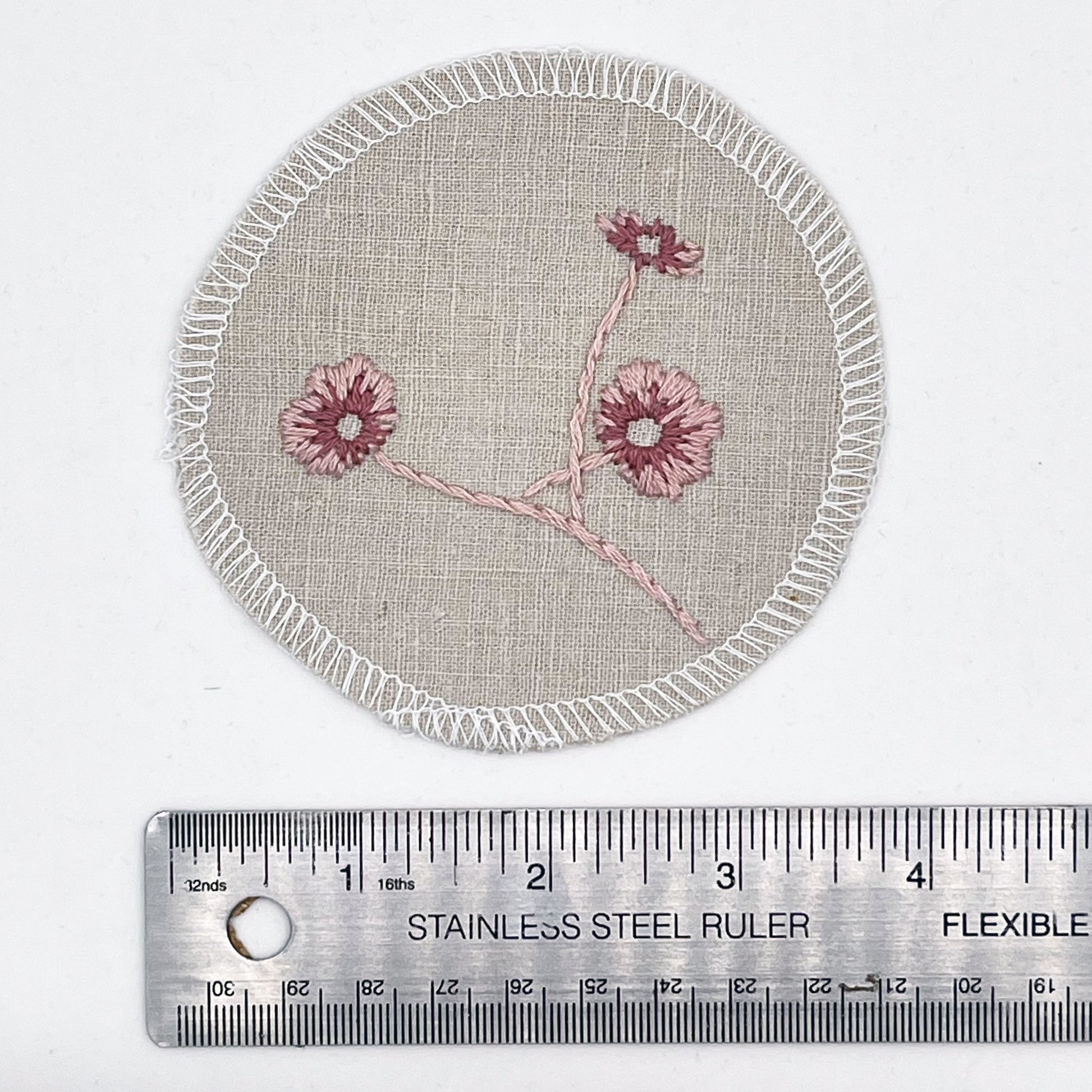 a patch made from a natural color cotton linen fabric placed next to a ruler, showing a diameter of 4 inches, hand embroidered with 3 poppies in shades of pink, edges overlocked with white thread, on a white background