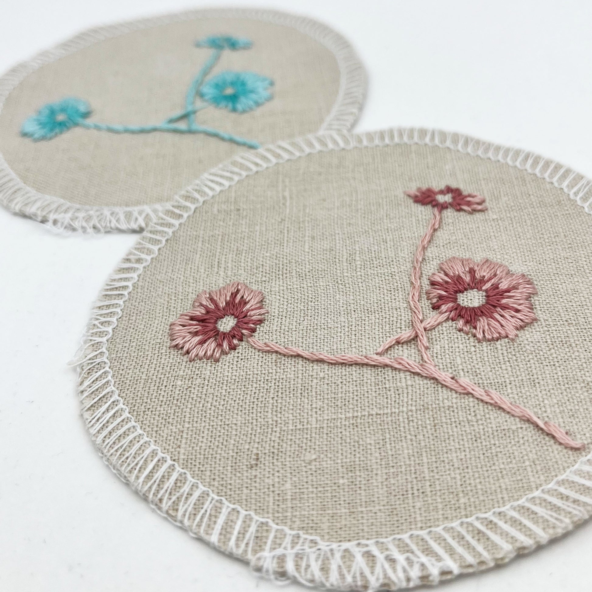Two circle shaped patches, made from a natural color linen fabric, hand embroidered with 3 poppies, one patch in pink and one in robin's egg blues, edges overlocked with white thread, on a white background