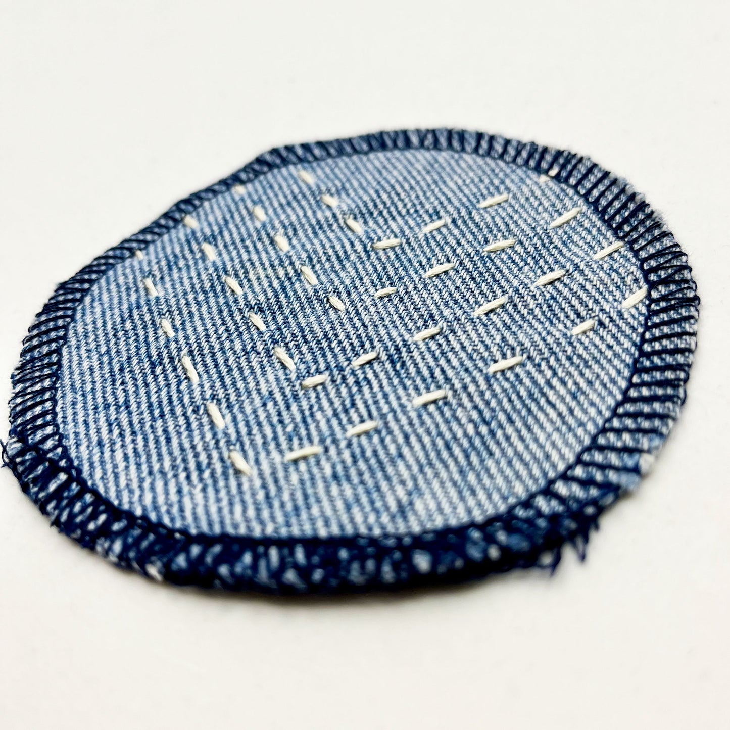 close up angled view of a circle shaped patch made out of denim, with overlocked edges, with rows of ivory running stitches in a chevron pattern, on a white background