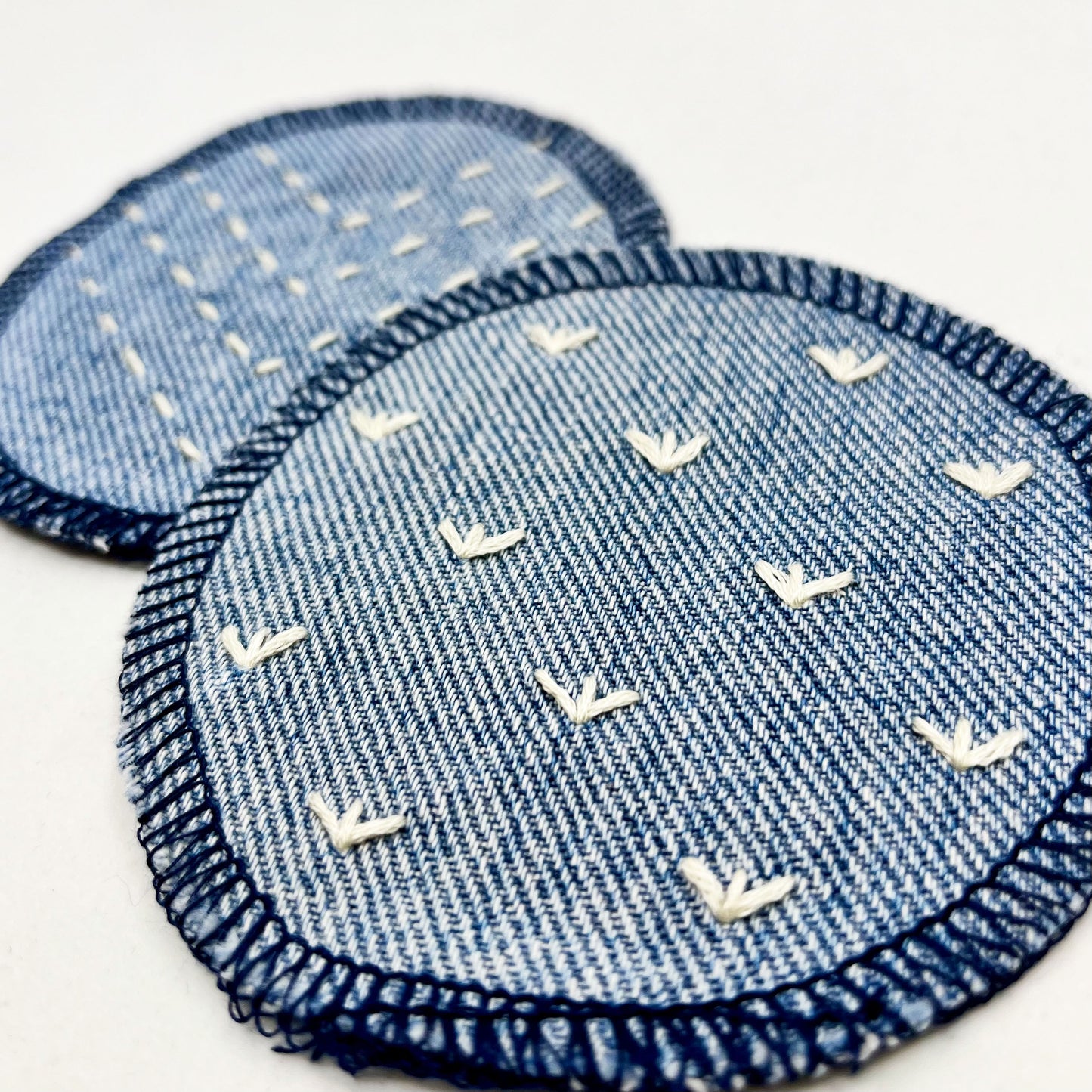 close up angled view of two circle shaped patches made out of denim, with overlocked edges, one with stitches in ivory that look like sprouts or birds feet, the other with rows of running stitches in a chevron pattern