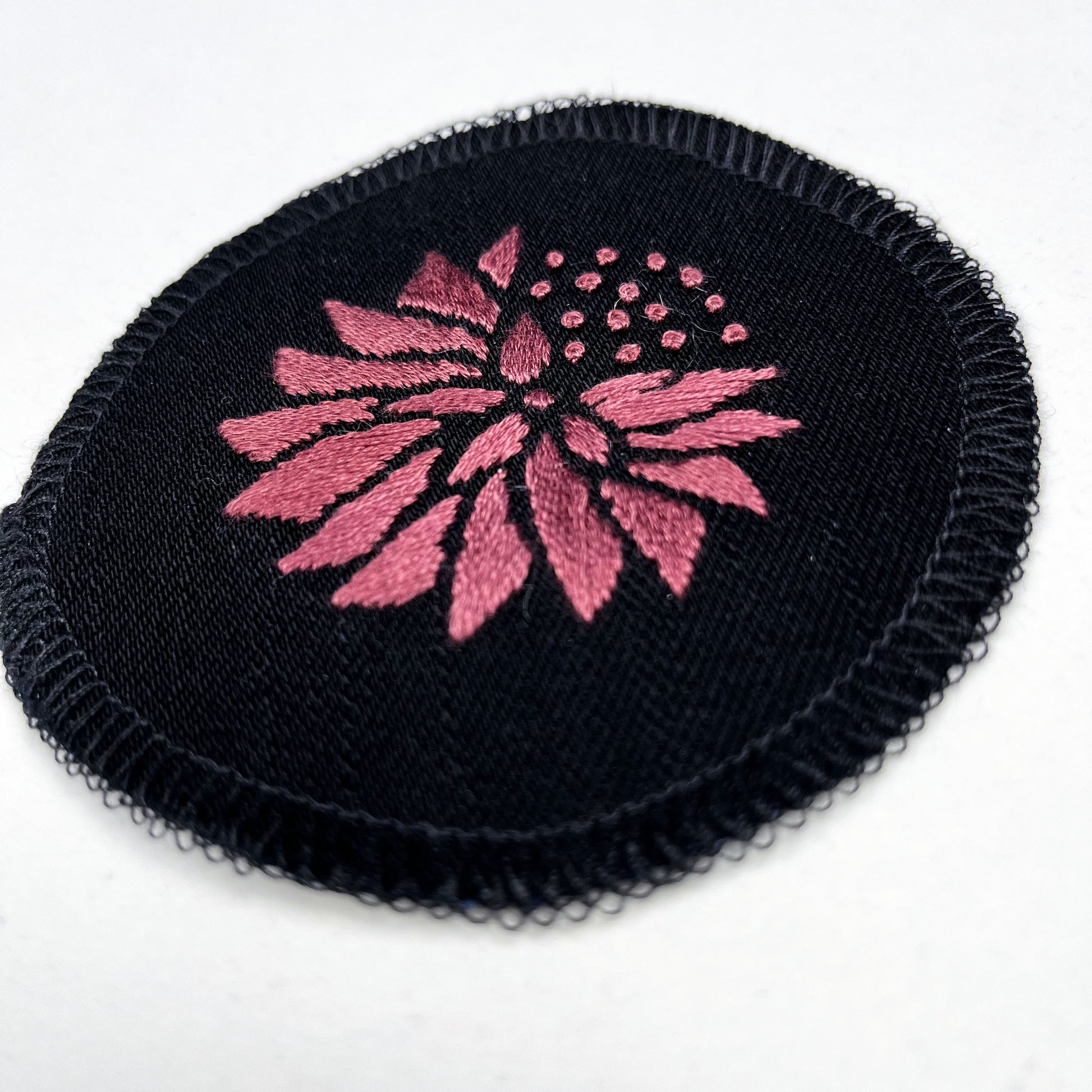 a circle shaped patch made out of black twill weave fabric, hand embroidered with an abstract dandelion in plum colored floss, with solid filled petals, edges overlocked in black, on a white background