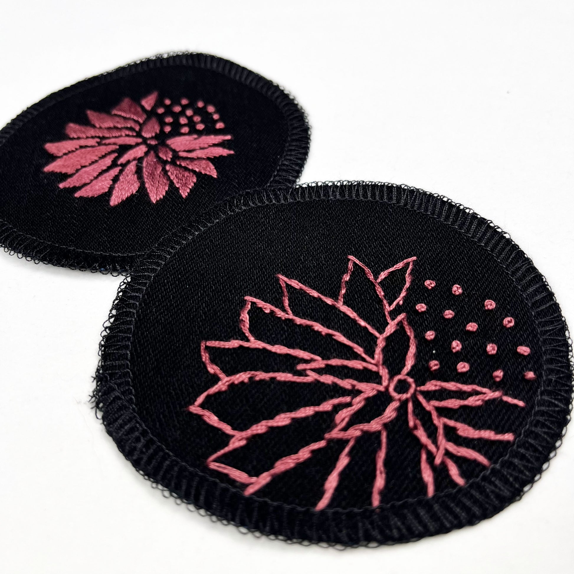 two circle shaped patches made out of black twill weave fabric, hand embroidered with abstract dandelions in plum colored floss, one with solid filled petals, the other with outlines of petals, edges overlocked in black, on a white background
