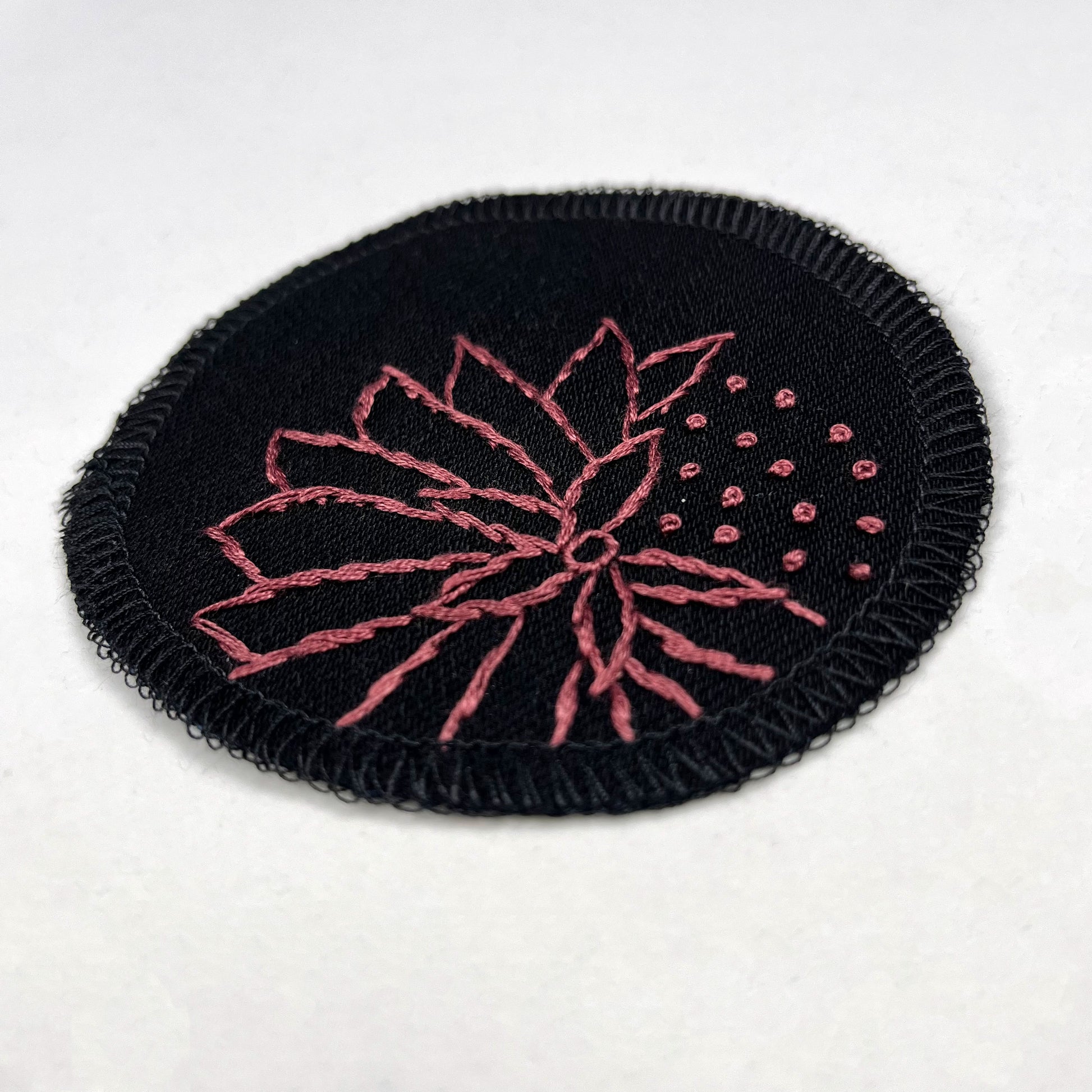 a circle shaped patch made out of black twill weave fabric, hand embroidered with an abstract dandelion in plum colored floss, outlines of petals, edges overlocked in black, on a white background