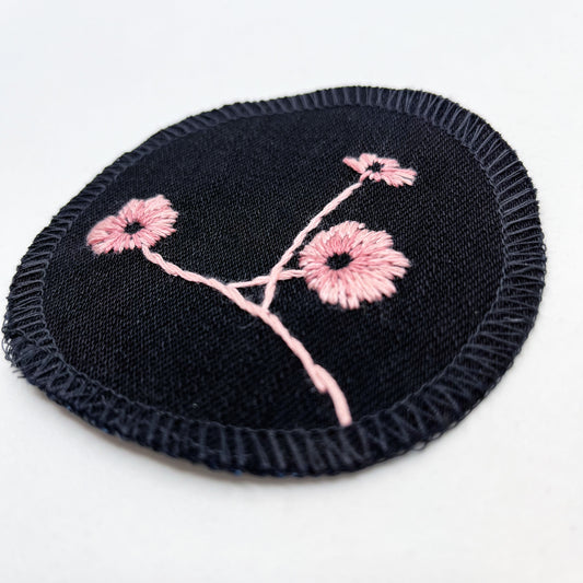 close up angled view of a circle shaped patch, made from black denim fabric, hand embroidered with 3 poppies in shades of pink, edges overlocked with black thread, on a white background