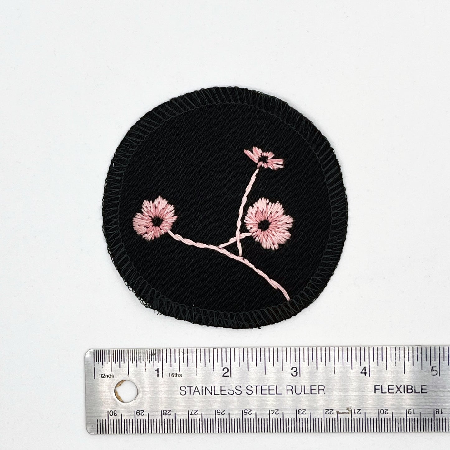 a circle shaped patch, made from black denim fabric, hand embroidered with 3 poppies in shades of pink, edges overlocked with black thread, placed next to a metal ruler to show a diameter of four inches, on a white background