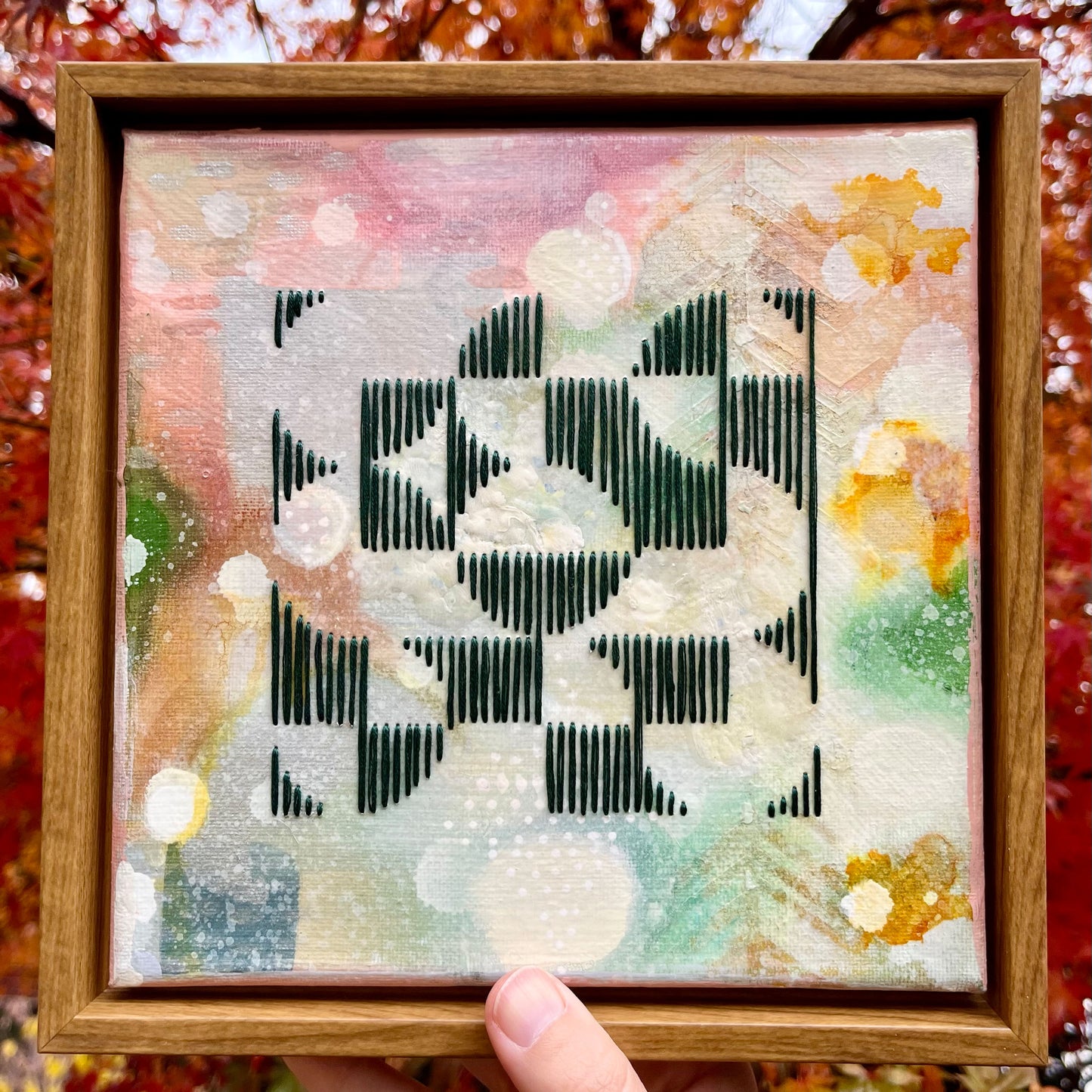 colorful square painting in a wood frame with an abstract image of divided circles in a grid hand stitched on it in emerald green embroidery floss, being held up with a background of red leaves on a tree