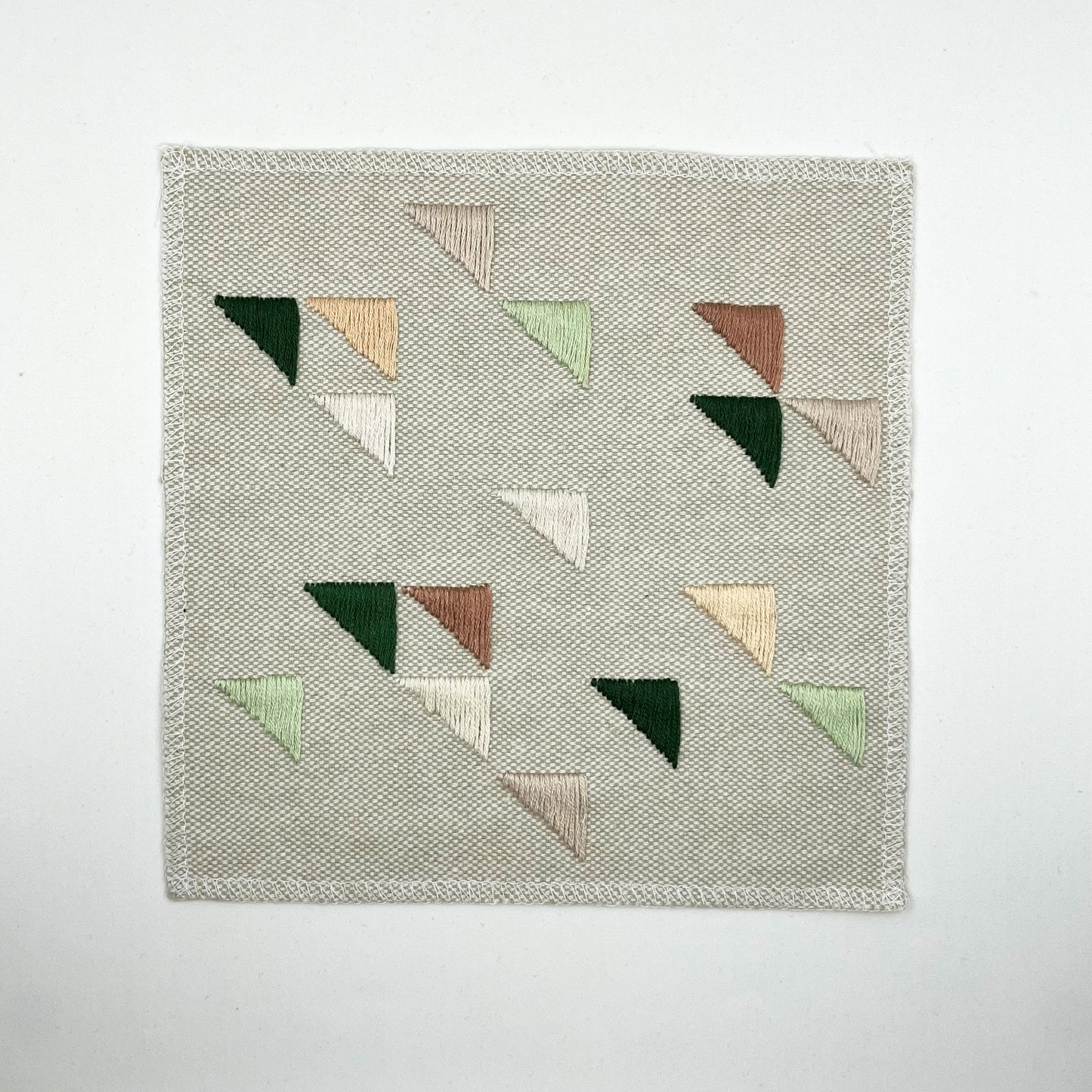 Large square natural colored patch embroidered with randomly placed triangles in peaches, mauves and greens, on a white background