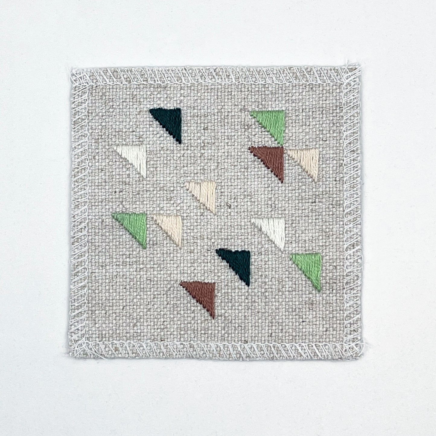 a square natural colored patch embroidered with randomly placed triangles in peaches, mauves and greens, on a white background