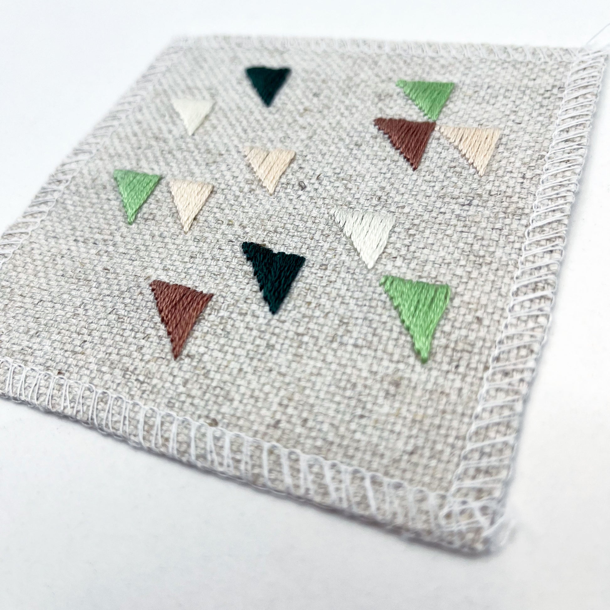 close up angled view of a square natural colored patch embroidered with randomly placed triangles in peaches, mauves and greens, on a white background