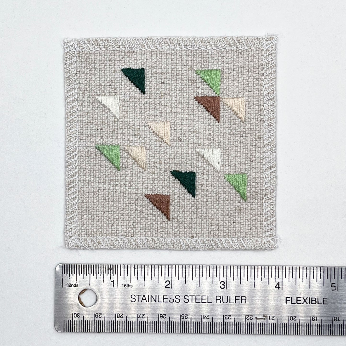 a square natural colored patch embroidered with randomly placed triangles in peaches, mauves and greens, placed next to a metal ruler to show a width of four inches, on a white background