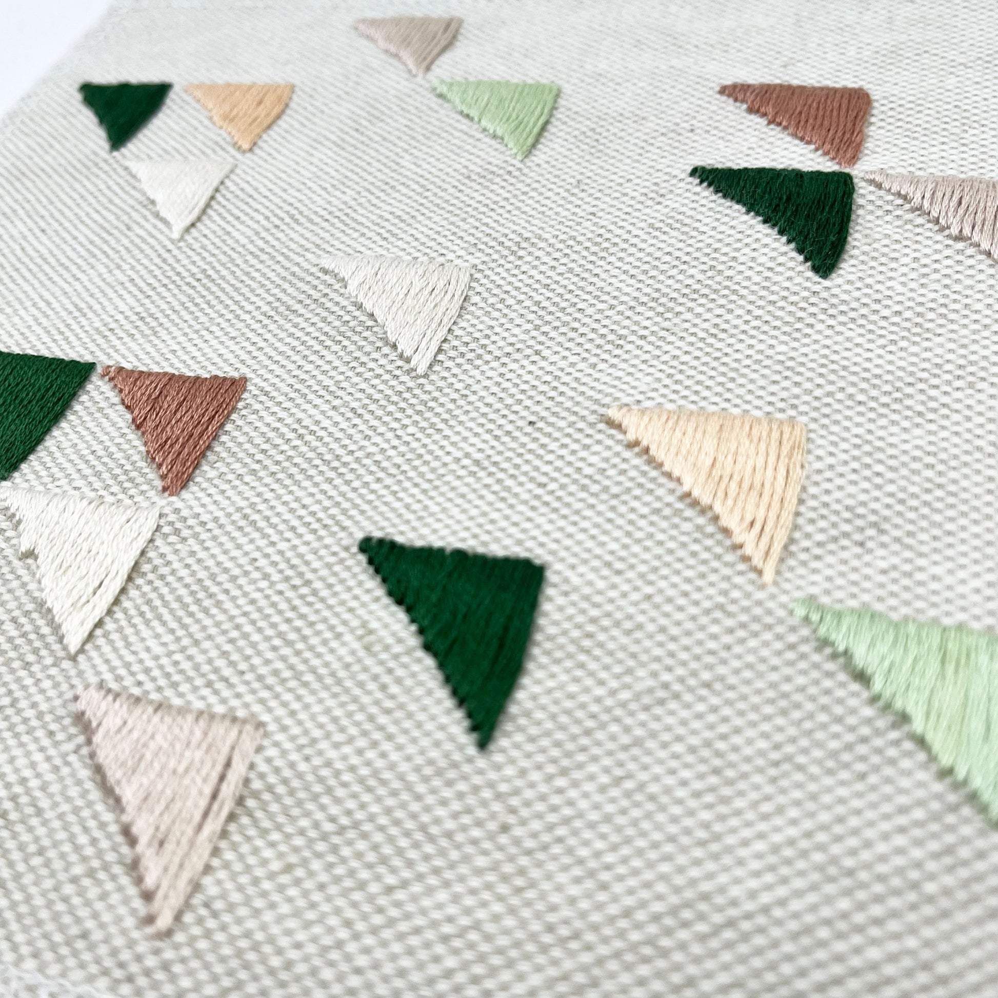 close up angled view of a large square natural colored patch embroidered with randomly placed triangles in peaches, mauves and greens