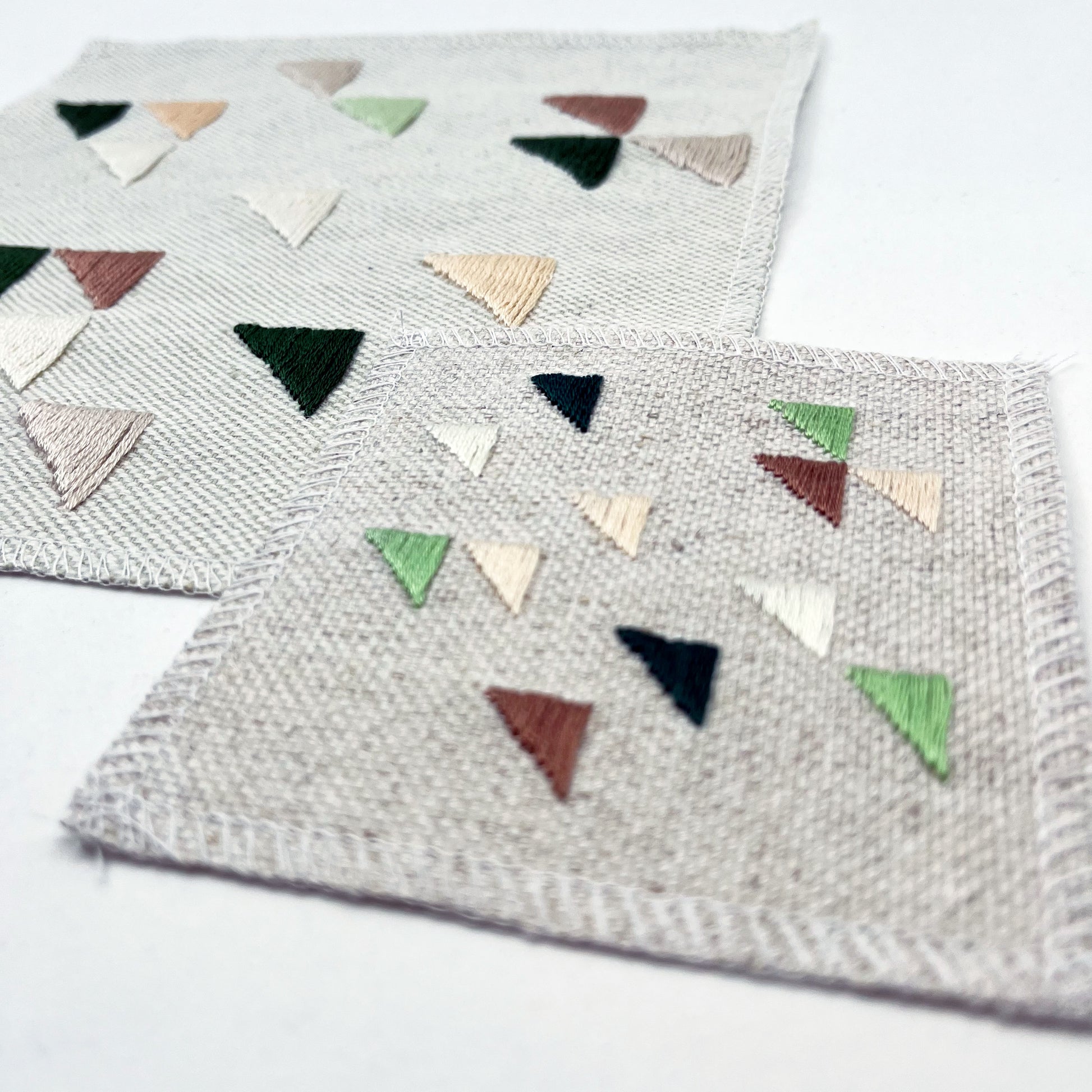 close up angled view of two square natural colored patches in different sizes embroidered with randomly placed triangles in peaches, mauves and greens, on a white background