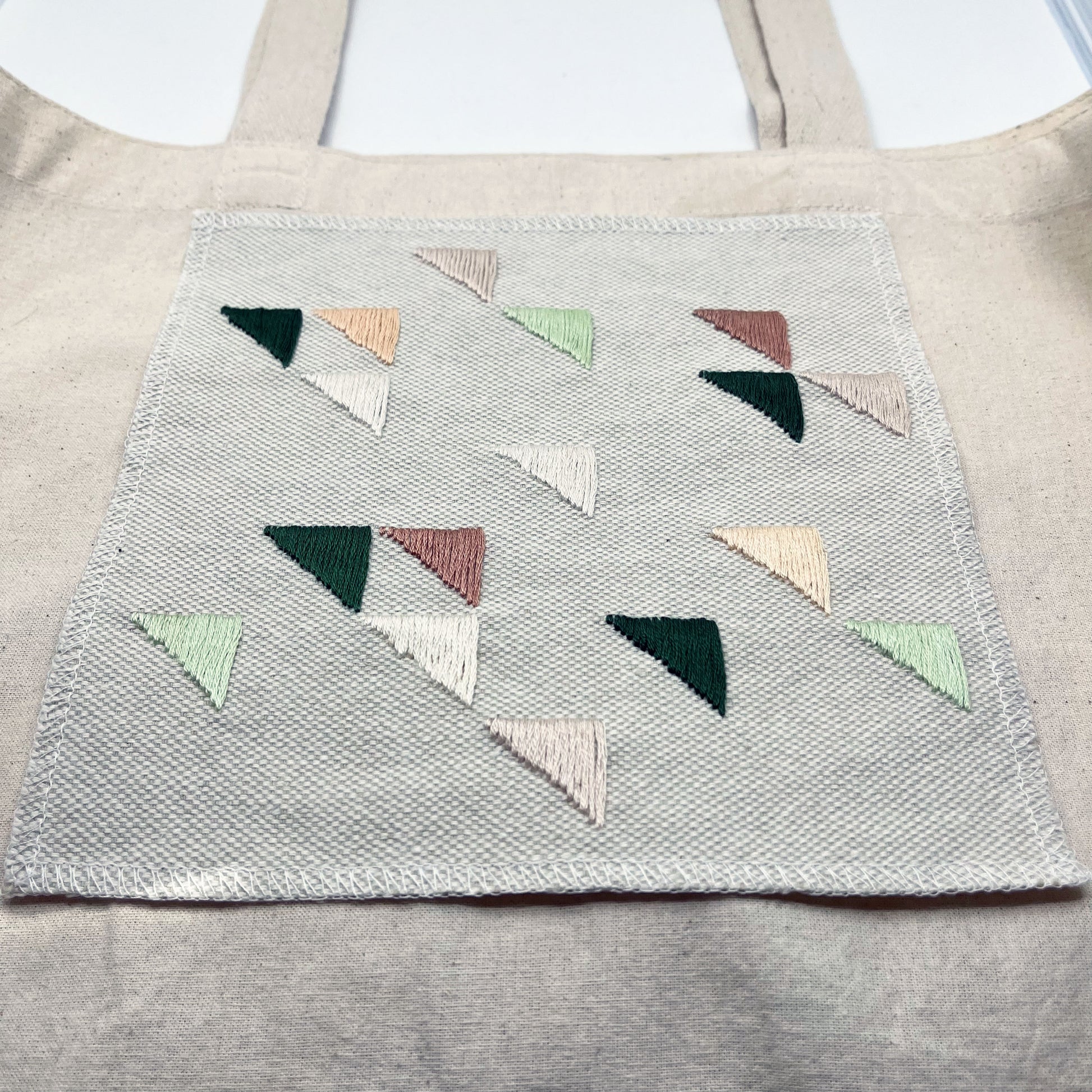 Large square natural colored patch embroidered with randomly placed triangles in peaches, mauves and greens, on a natural colored canvas tote bag